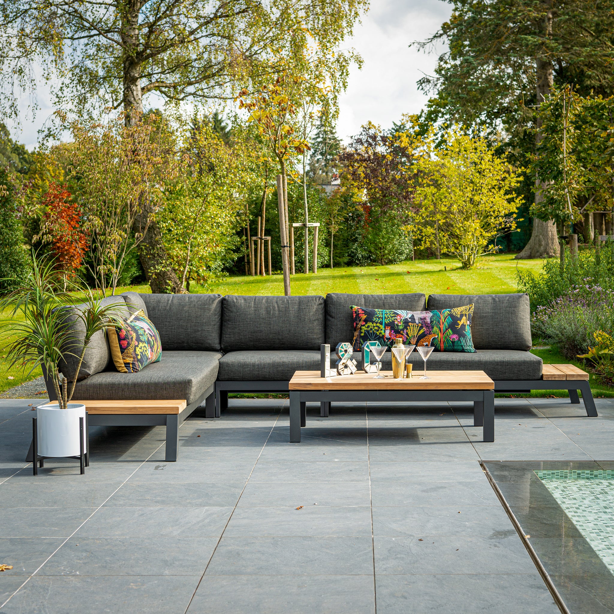 The Empire Large Corner Group Set with Teak Tables in Grey features a gray sectional sofa with olefin fabric, colorful pillows, and a wooden coffee table on tiled surfaces near a pool, all part of an elegant modular lounge series.