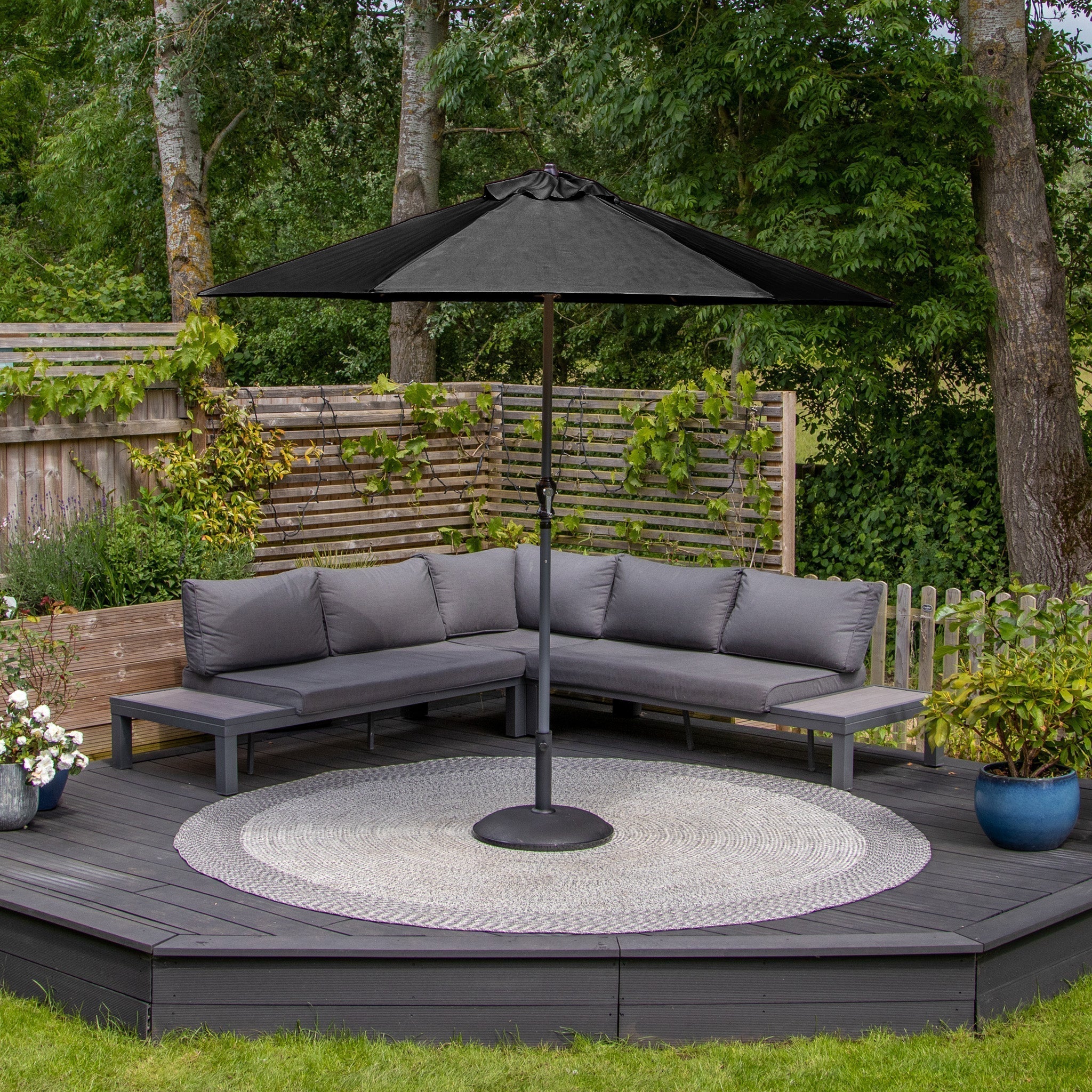 Modern outdoor seating featuring a grey sectional sofa, a fade-resistant Triton 2.5m Round Aluminium Parasol in Black with UV rating 50+, a circular rug, and greenery in the background.