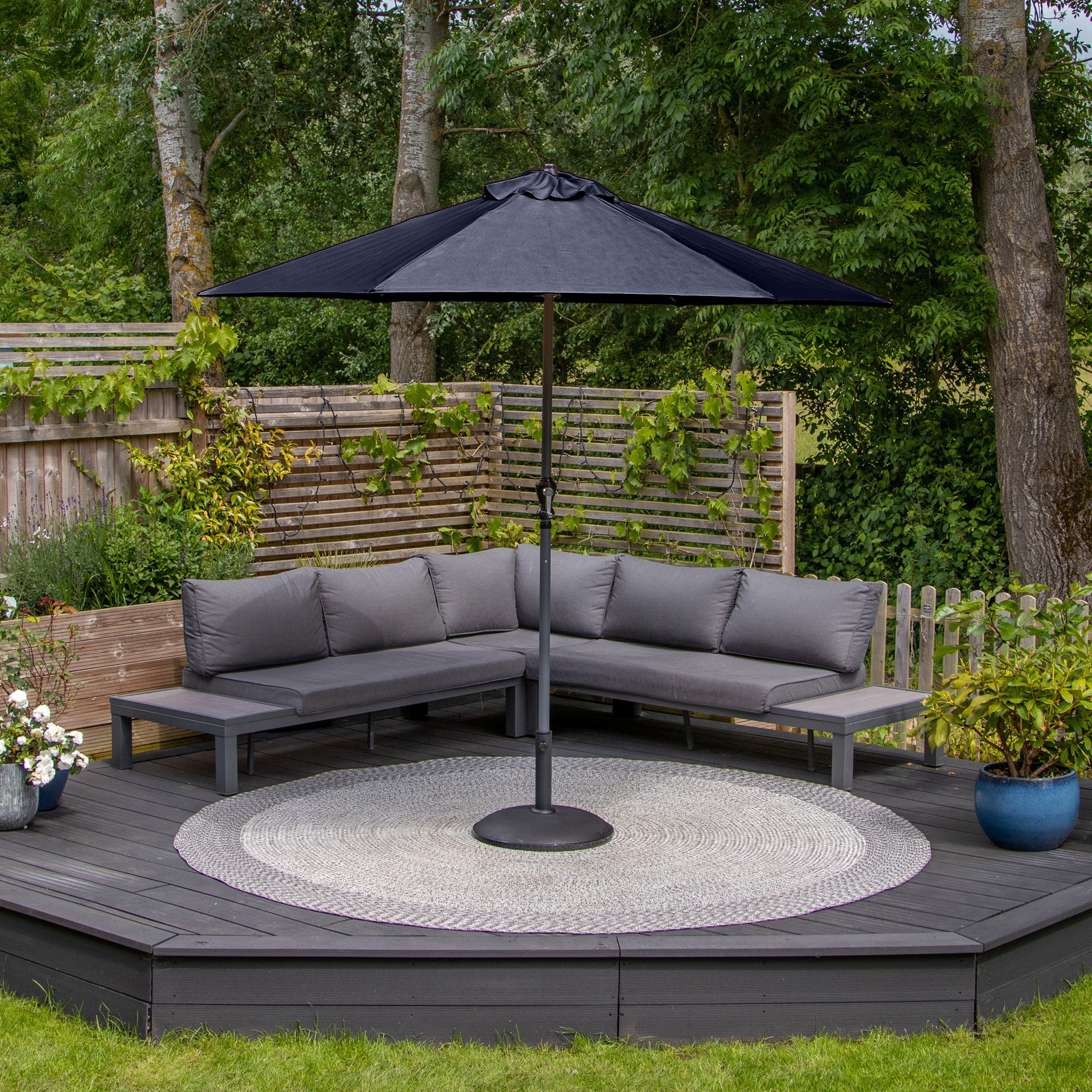 Outdoor seating area with gray sectional sofa, Triton 2.5m Round Aluminium Parasol in Navy featuring fade-resistant fabric and a UV rating of 50+, circular rug, and wooden deck, surrounded by greenery.