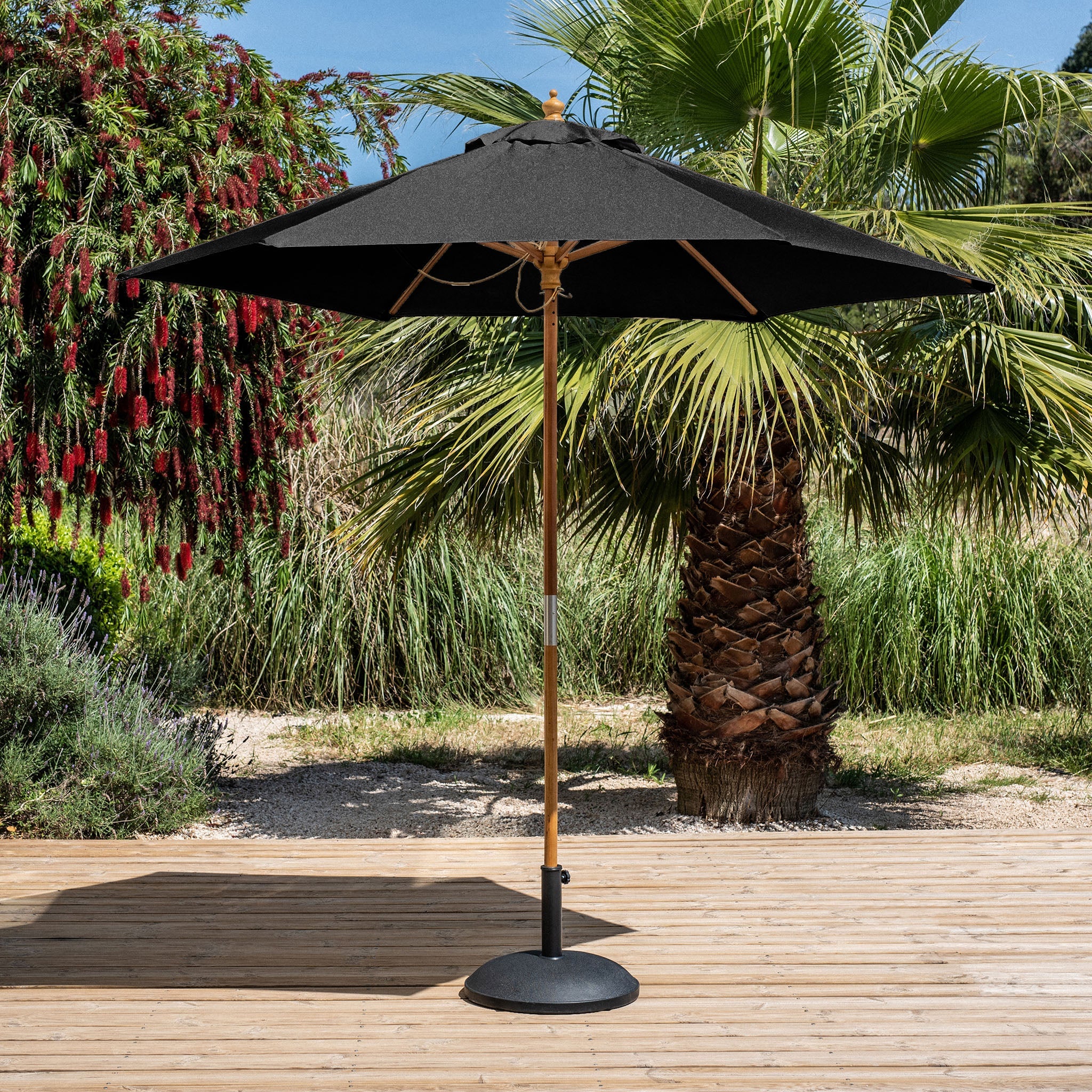 The Kratos 2.5m Round Wooden Parasol in Black showcases its UV-protected polyester canopy, elegantly set on a wooden pole amid a wooden deck, encircled by vibrant greenery and palm trees.