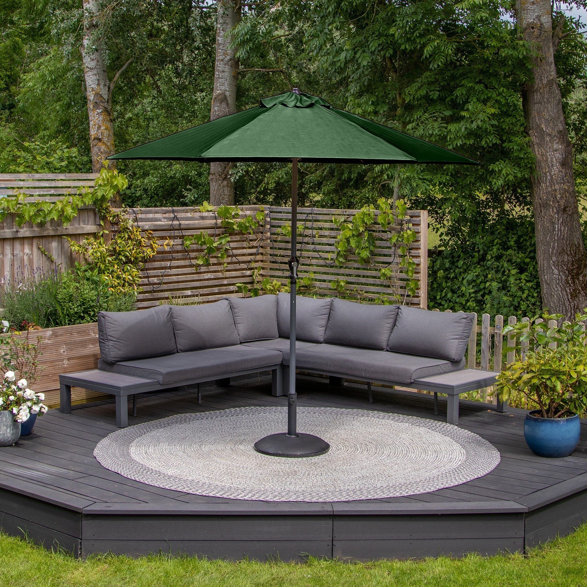 Outdoor patio with gray sectional sofa, round coffee table, Triton 2.5m Round Aluminium Parasol in Green with a UV rating of 50+, and wooden privacy screen with plants.