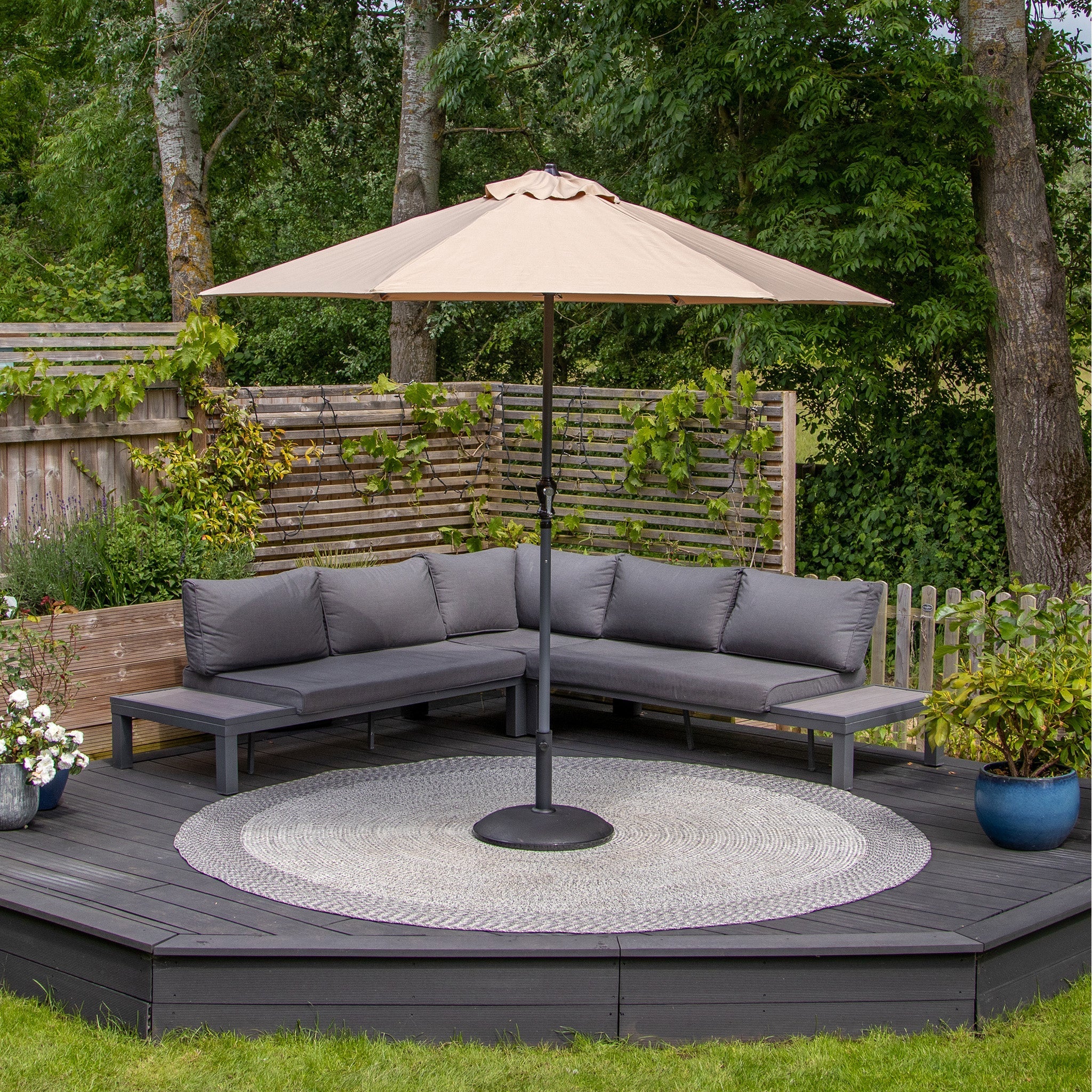 Outdoor seating area with a gray sectional sofa, khaki Triton 2.5m Round Aluminium Parasol, and circular rug on a wooden deck surrounded by greenery. The parasol offers a UV rating of 50+ and fade resistant fabric for lasting quality.