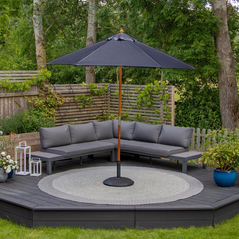 Gray outdoor patio set with sectional sofa, rug, and decorative plants on a raised wooden deck featuring the Kratos 2.5m Round Wooden Parasol in Navy with UV rating of 50+ and an easy-to-use pulley system for effortless adjustment.