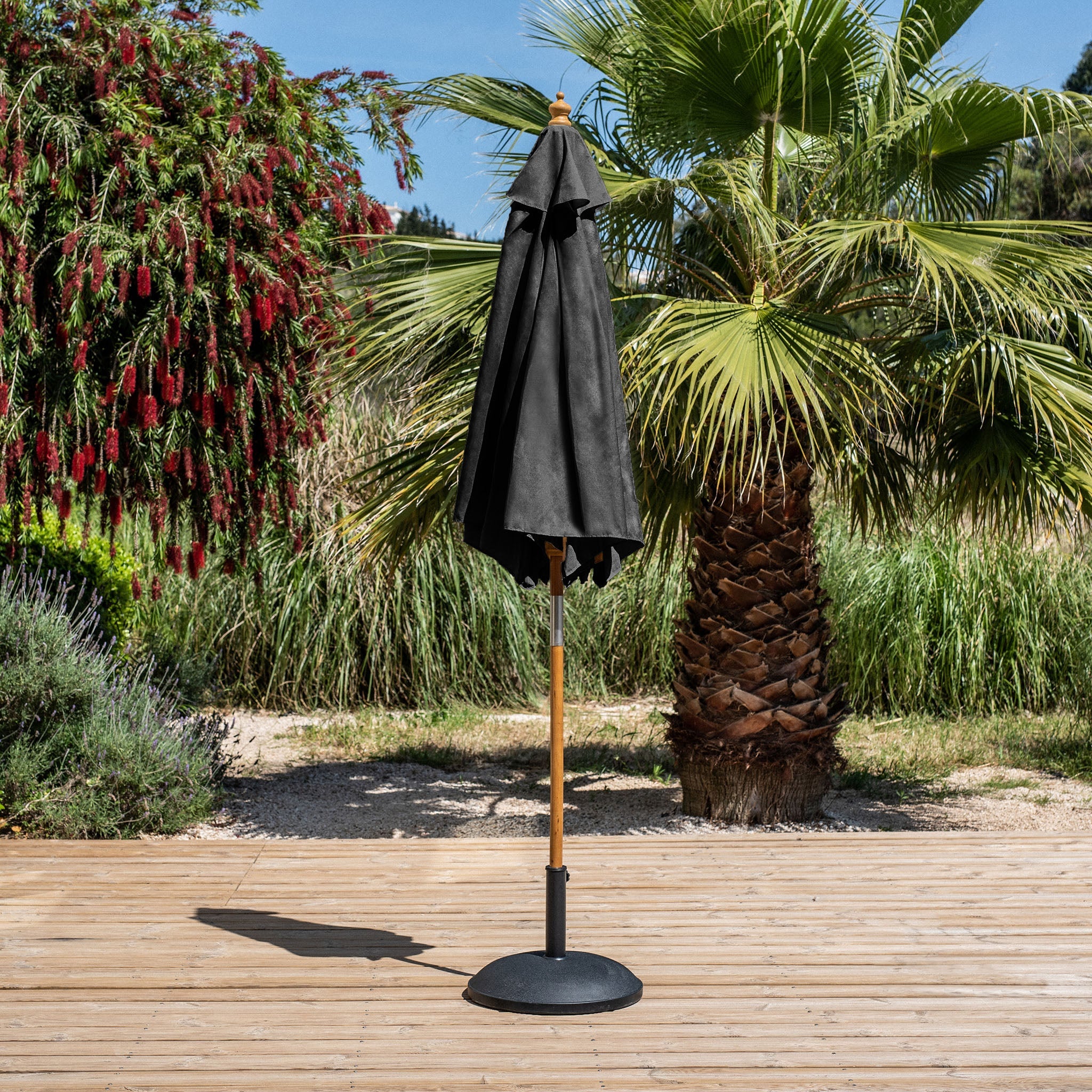 The Kratos 2.5m Round Wooden Parasol in Black is closed on a wooden deck, with lush green trees and plants in the background, showcasing its durable polyester canopy that offers excellent UV protection.