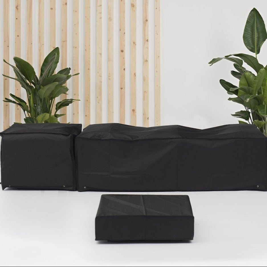 The Harbour Lifestyle Panama Luxury Outdoor Large Corner Set sits elegantly on a white patio, with tall green plants on both ends. The custom-fitted Winter Cover For Harbour Lifestyle Panama Luxury Outdoor Large Corner Set protects the black sofa and ottoman, ensuring durability and style throughout the seasons.