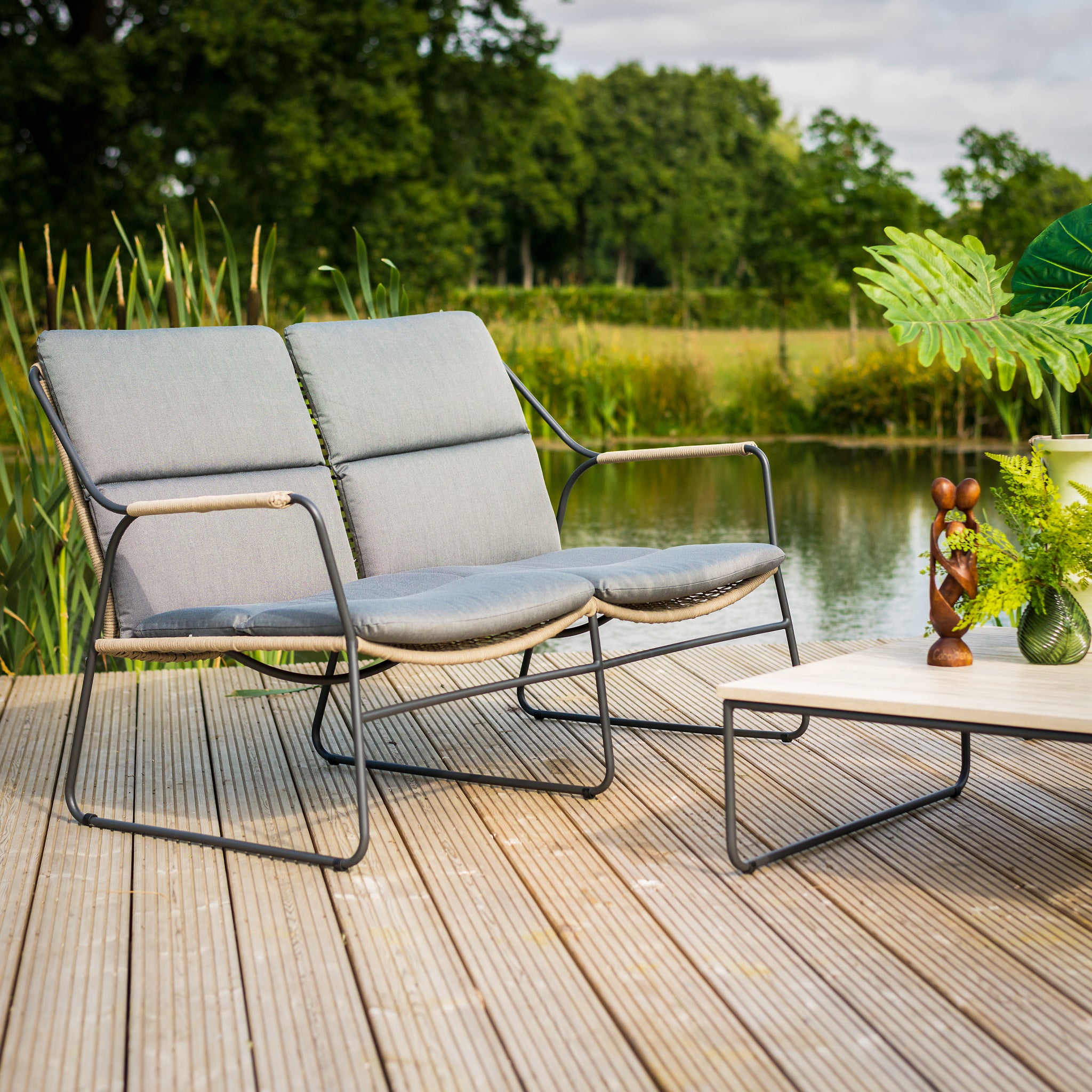 The Scandic 2 Seat Sofa Set with Teak Tables in Natural offers a garden furniture vibe on a wooden deck by a pond. Featuring a two-seater chair and table, it includes Olefin all-weather cushions, inviting you to relax among thriving plants and greenery.