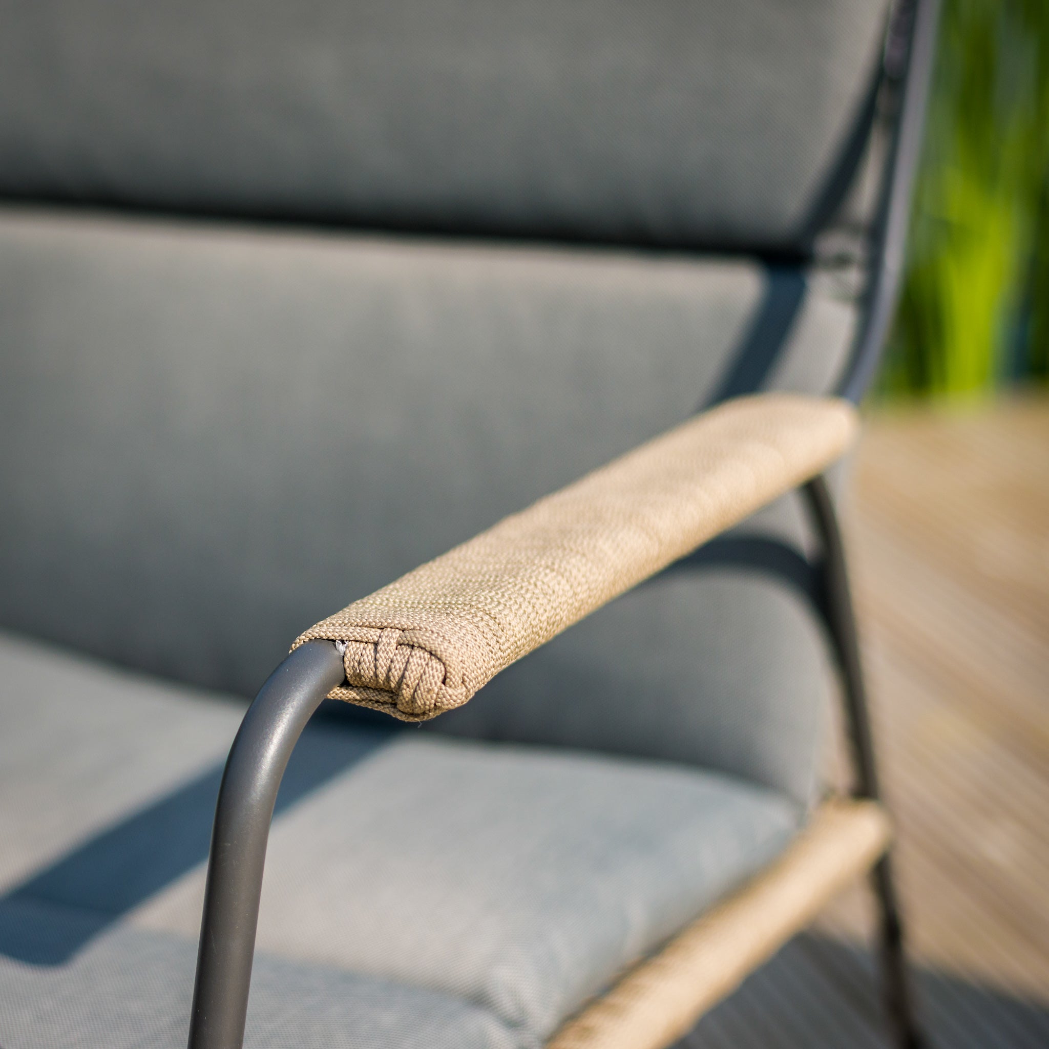 Close-up of a beige cushioned armrest on a gray outdoor chair with Olefin all-weather cushions, set against a blurred wooden deck and greenery, showcasing the elegance of the Scandic 2 Seat Sofa Set with Teak Tables in Natural.