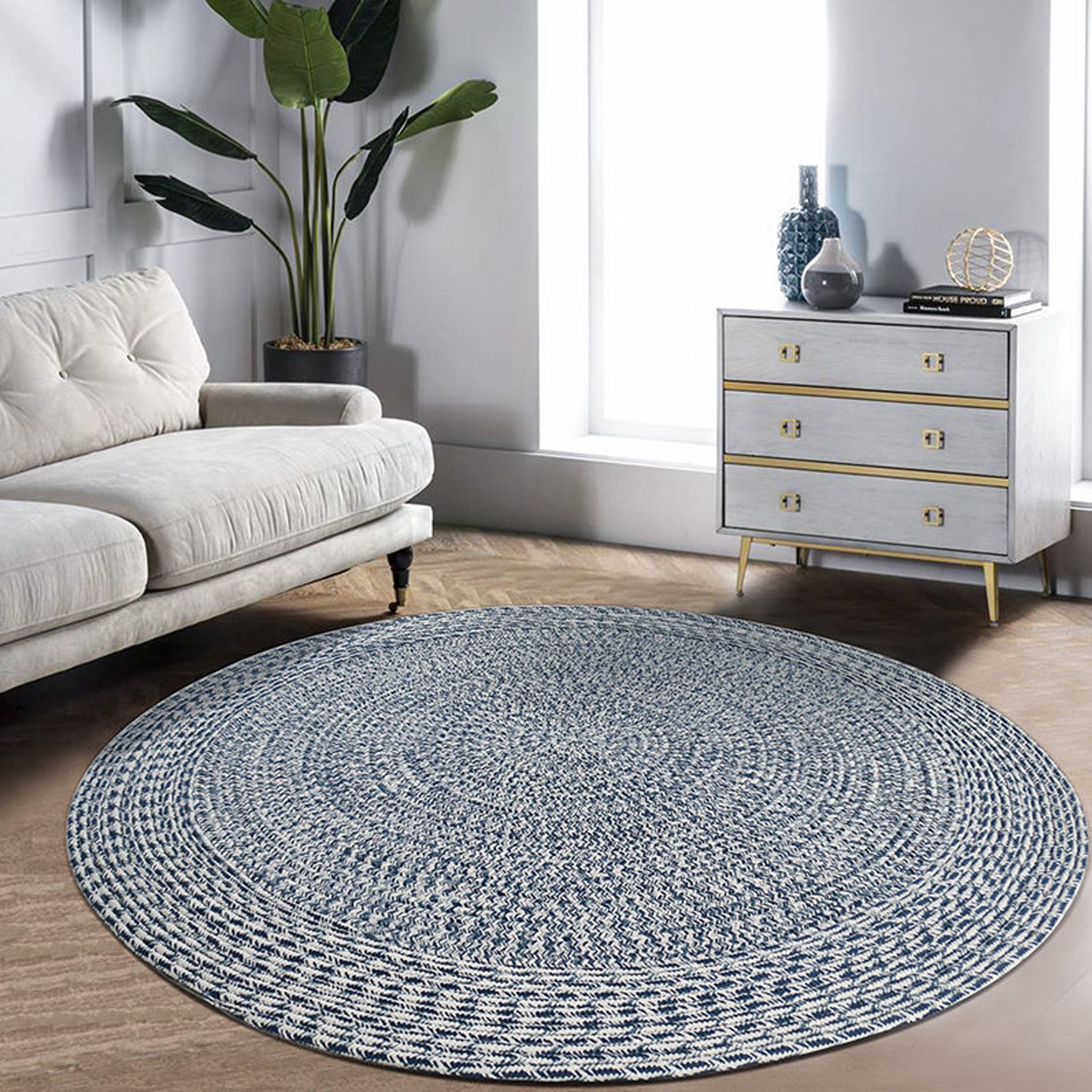 Apollo 300cm Round Indoor and Outdoor Rug in Prussian Blue in a modern living room with a light grey sofa, plants, and a white chest of drawers by a large window. This UV-resistant, weatherproof rug brings both style and durability to your space.