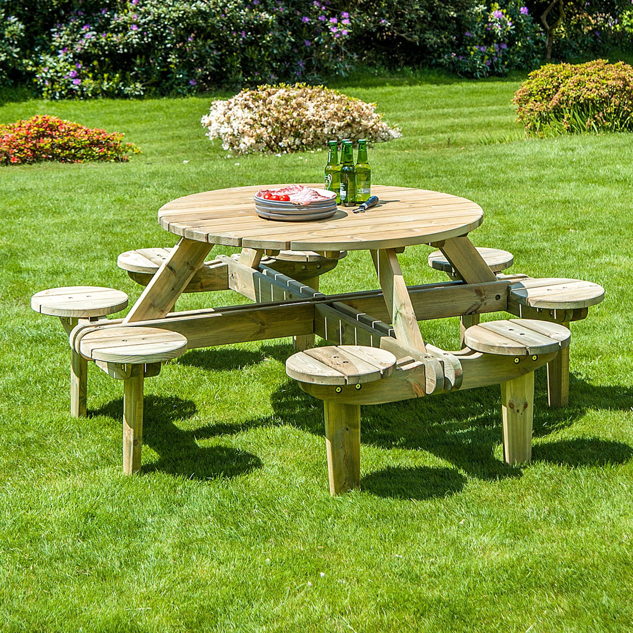 The Pine Wood Gleneagles 8 Seater Round Picnic Table, designed with attached stools and crafted from pine sourced from responsibly managed forests, hosts a few bottles and dishes. It is set on a lush green lawn, perfect for outdoor use.