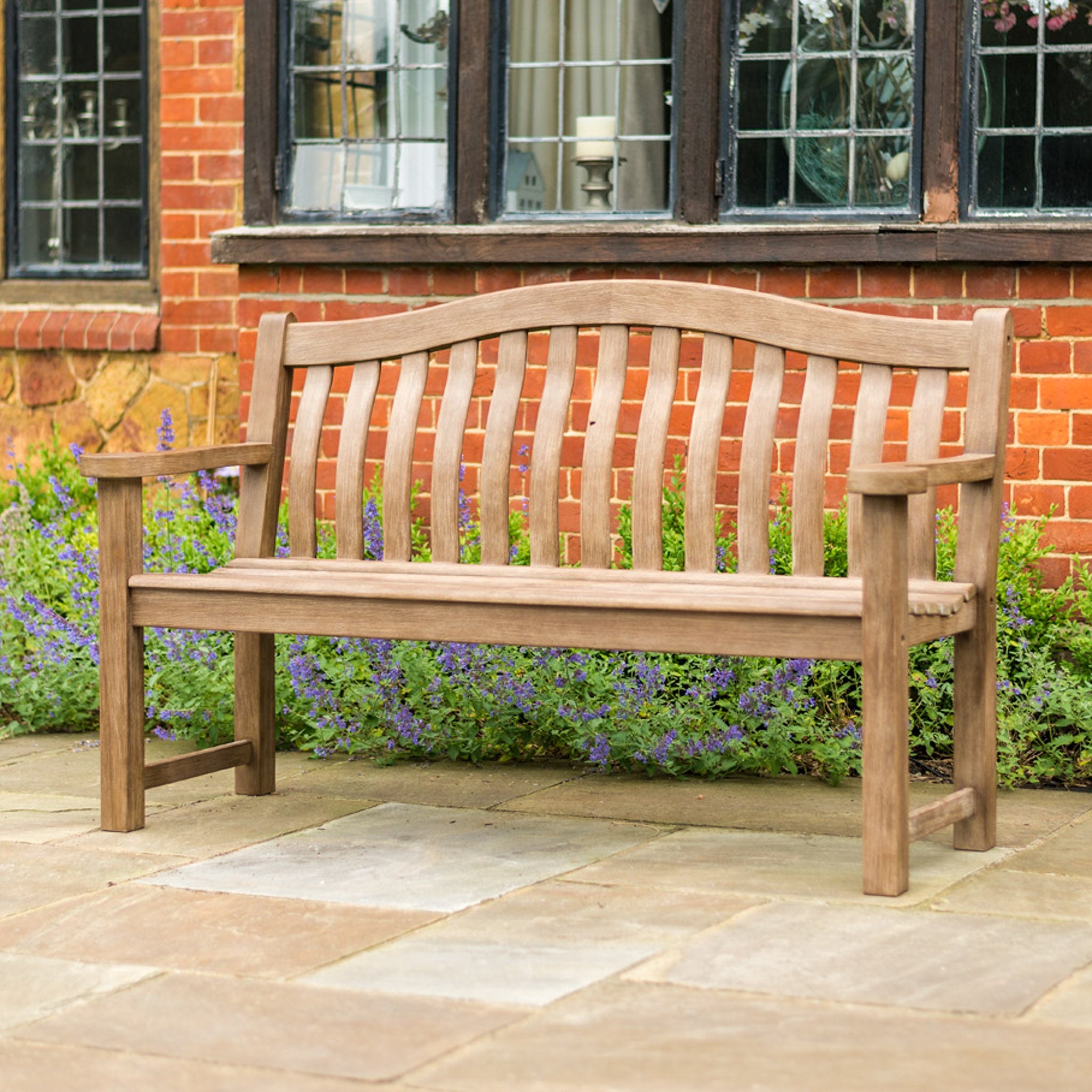 A Sherwood Acacia Turnberry Bench - 5ft on a patio in front of a brick building with windows and flowering plants in the background.