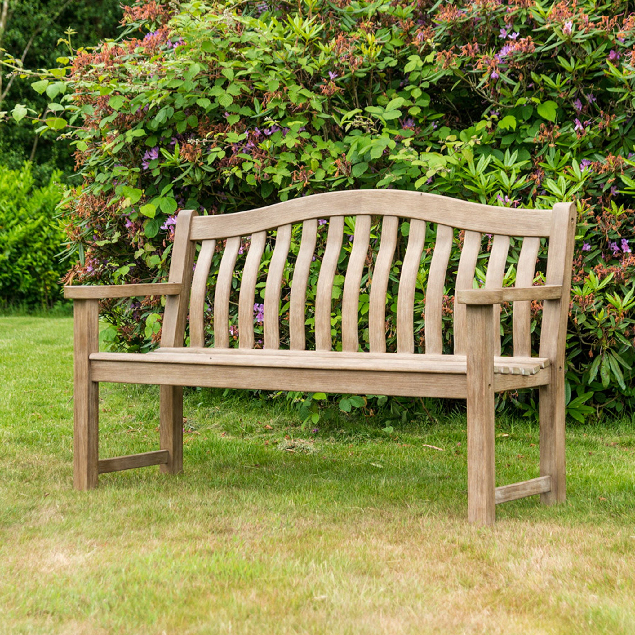 A Sherwood Acacia Turnberry Bench - 5ft, crafted from acacia timber, sits gracefully on the grass, surrounded by lush greenery and flowering bushes in the background.