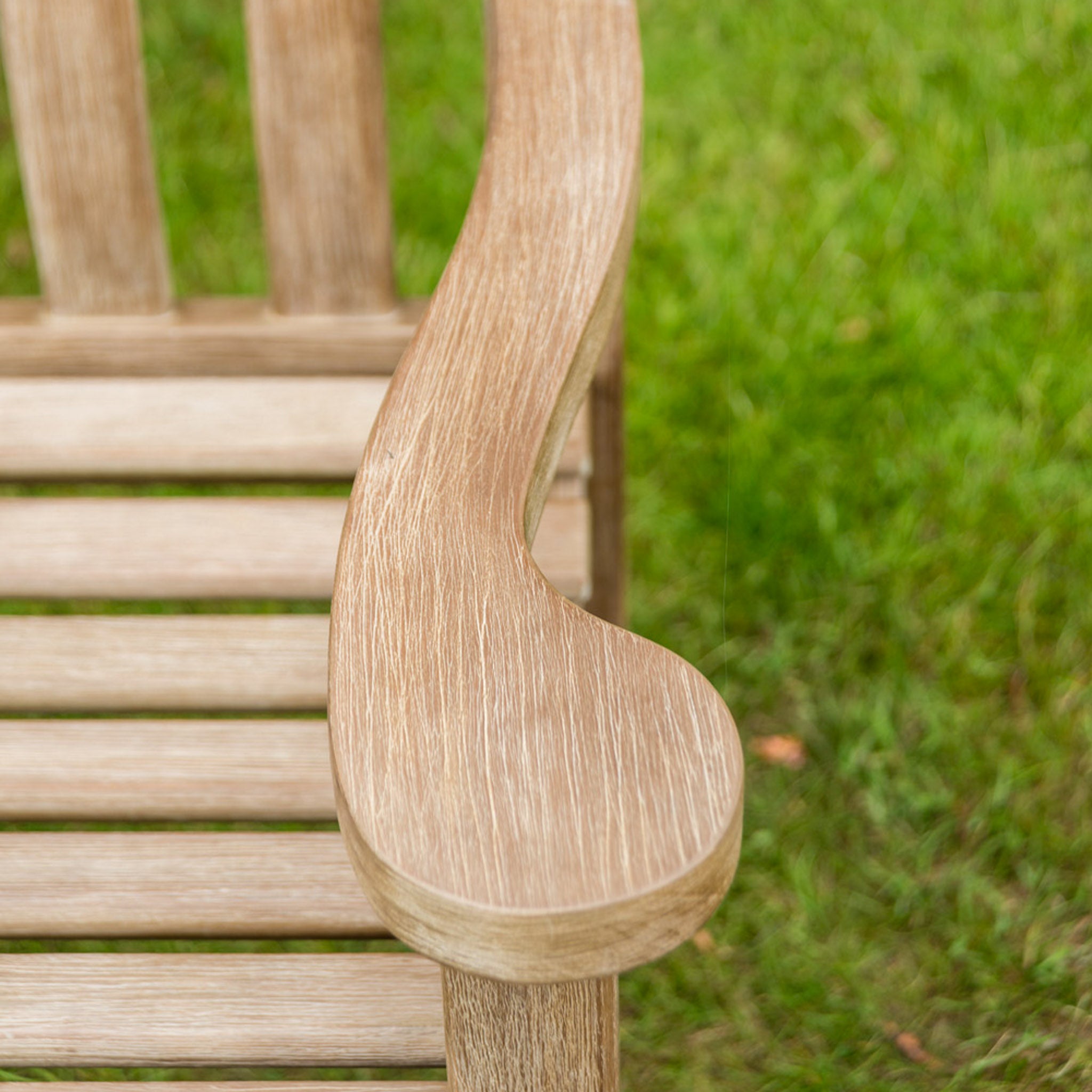 Close-up of a wooden armrest on a Sherwood Acacia Turnberry Bench - 5ft, with green grass in the background.