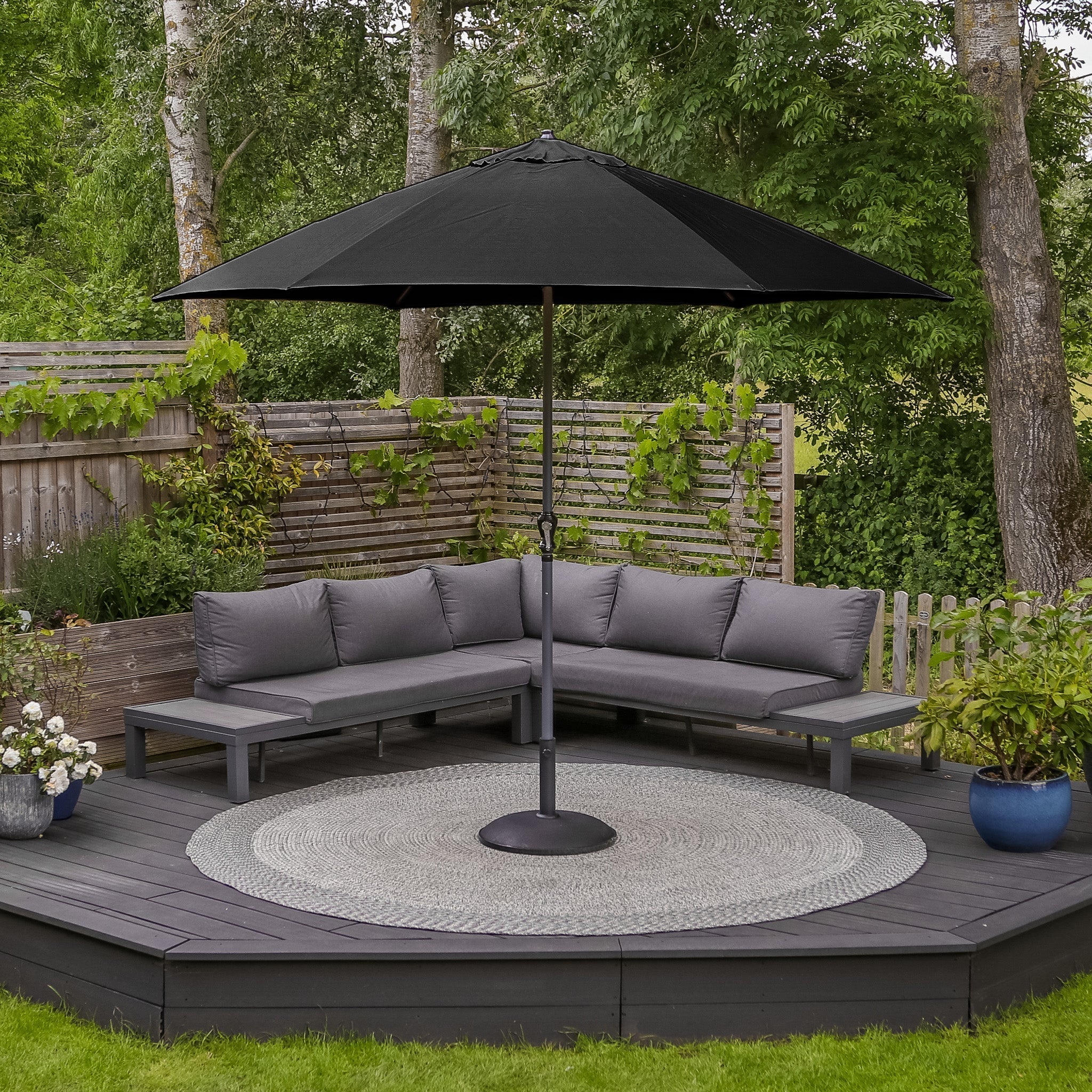 Outdoor patio with a gray sectional sofa, the Triton 3m Round Aluminium Parasol in Black featuring a crank handle system and UV rating of 50+, complemented by a circular rug on the wooden deck, surrounded by greenery.