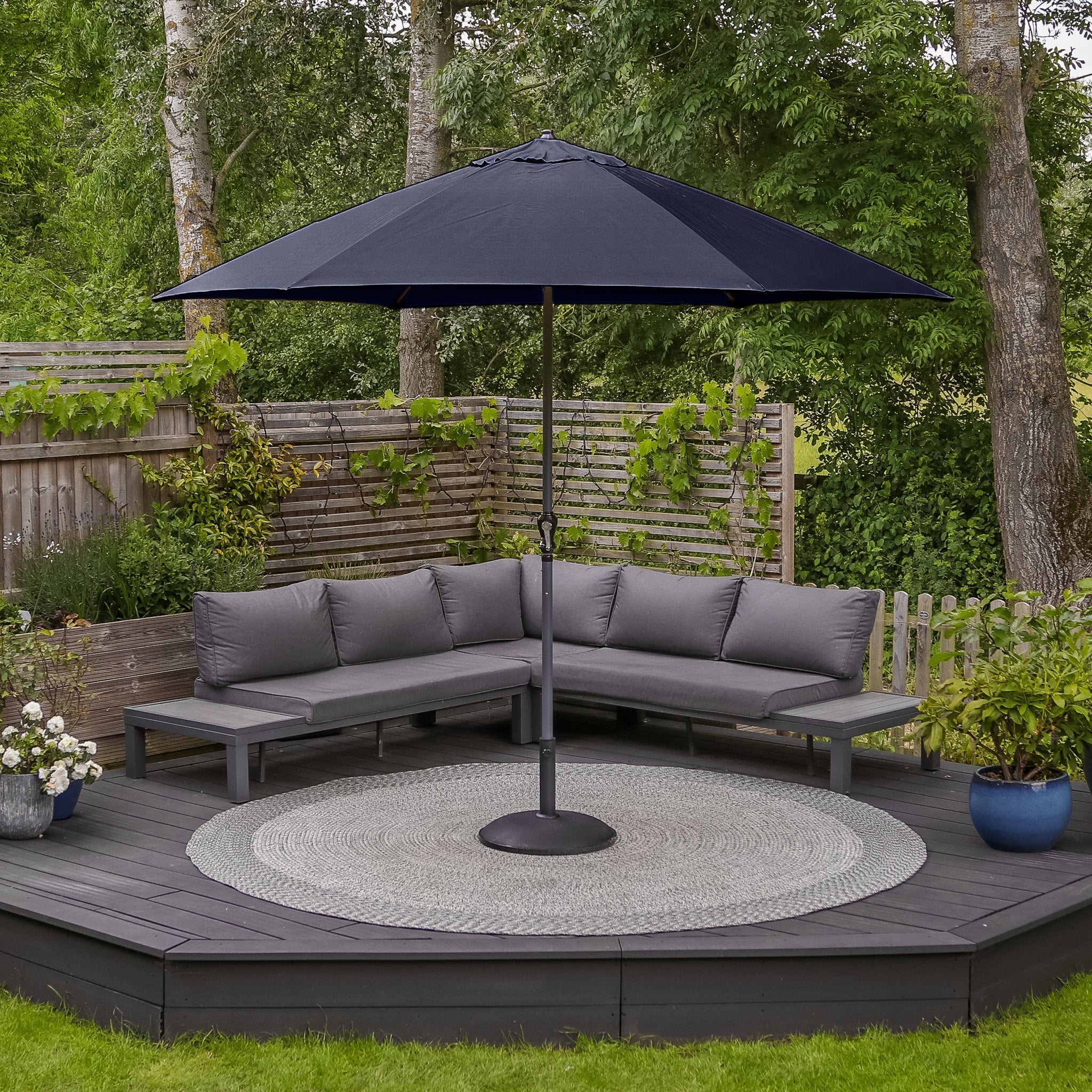 A patio featuring a gray sectional sofa, circular rug, and the impressive 3-meter Triton Round Aluminium Parasol in Navy with a UV rating of 50+, all set against a backdrop of lush greenery.