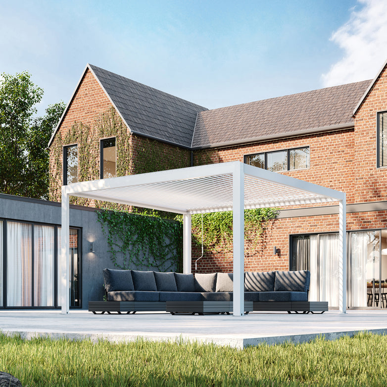 A modern patio showcases a white PergoSTET Plus pergola with tilting louvres and LED lighting, alongside a gray sectional sofa, set against a brick house under the blue sky.