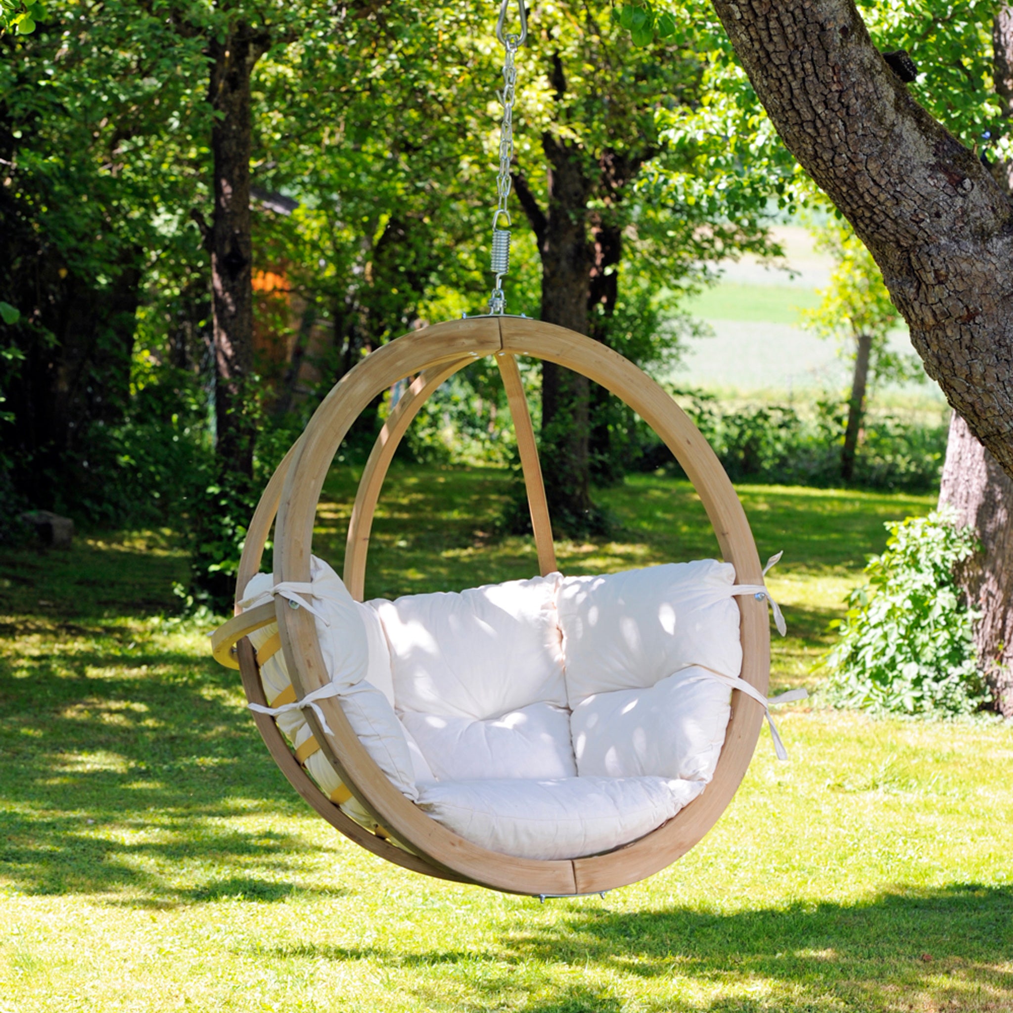 The Natura Globo Single Hanging Chair, featuring elegant white cushions, is suspended from a tree in a lush green garden, making it the perfect addition to both indoor and outdoor living spaces.