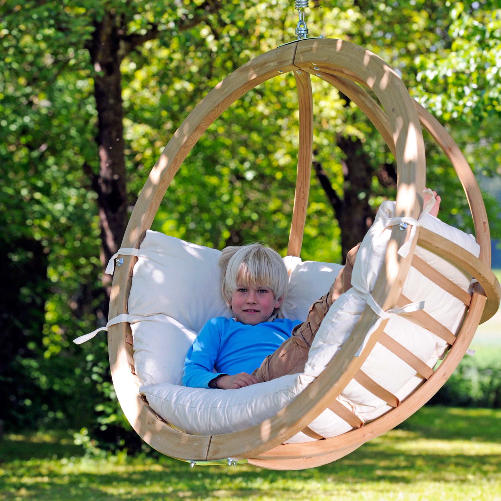A child with blond hair relaxes in a round, wooden Natura Globo Single Hanging Chair outdoors on a sunny day, surrounded by green trees. This stylish furniture piece seamlessly integrates comfort into both indoor and outdoor living spaces.