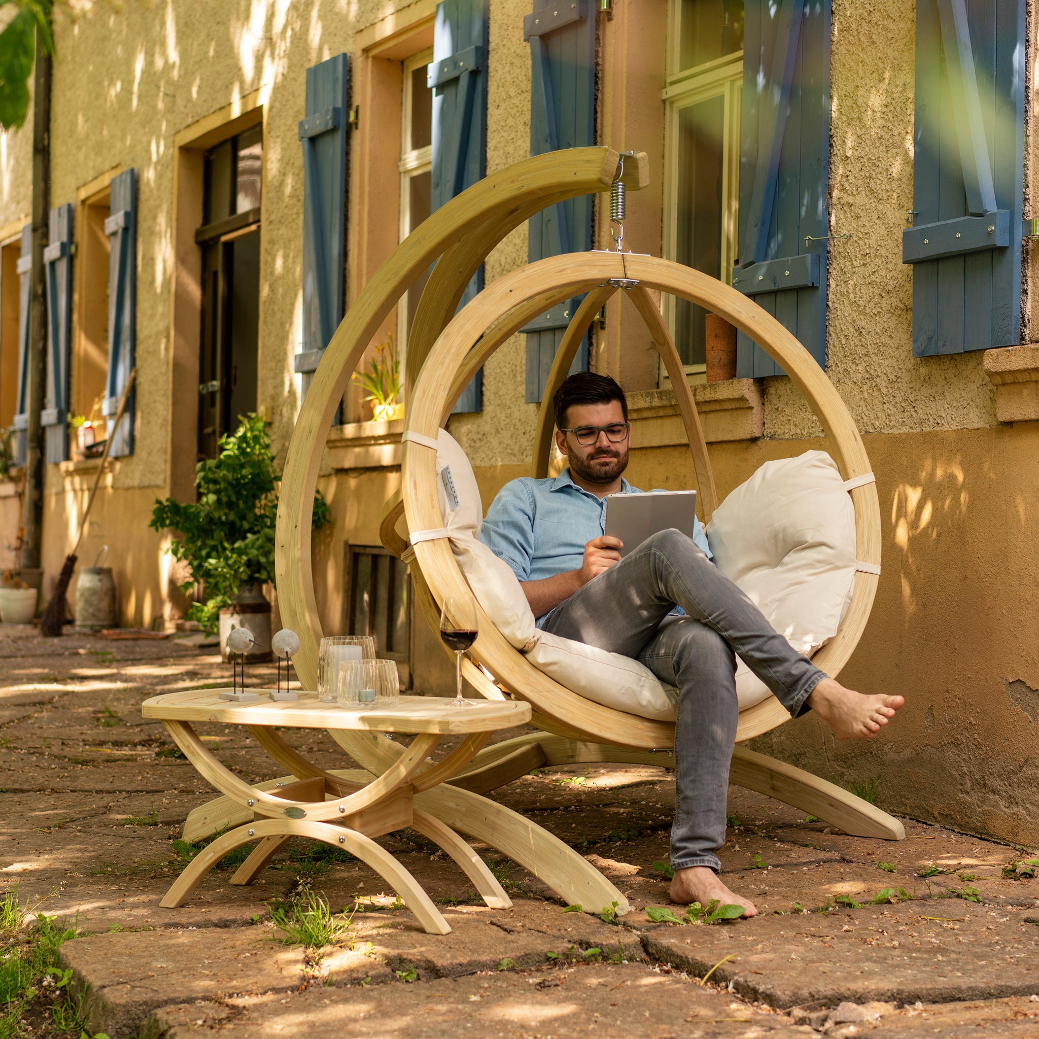 A man sits comfortably in the Natura Globo Single Hanging Chair on a rustic patio, reading a book beside a round wooden table adorned with decor items, seamlessly blending indoor and outdoor living spaces with stylish furniture.