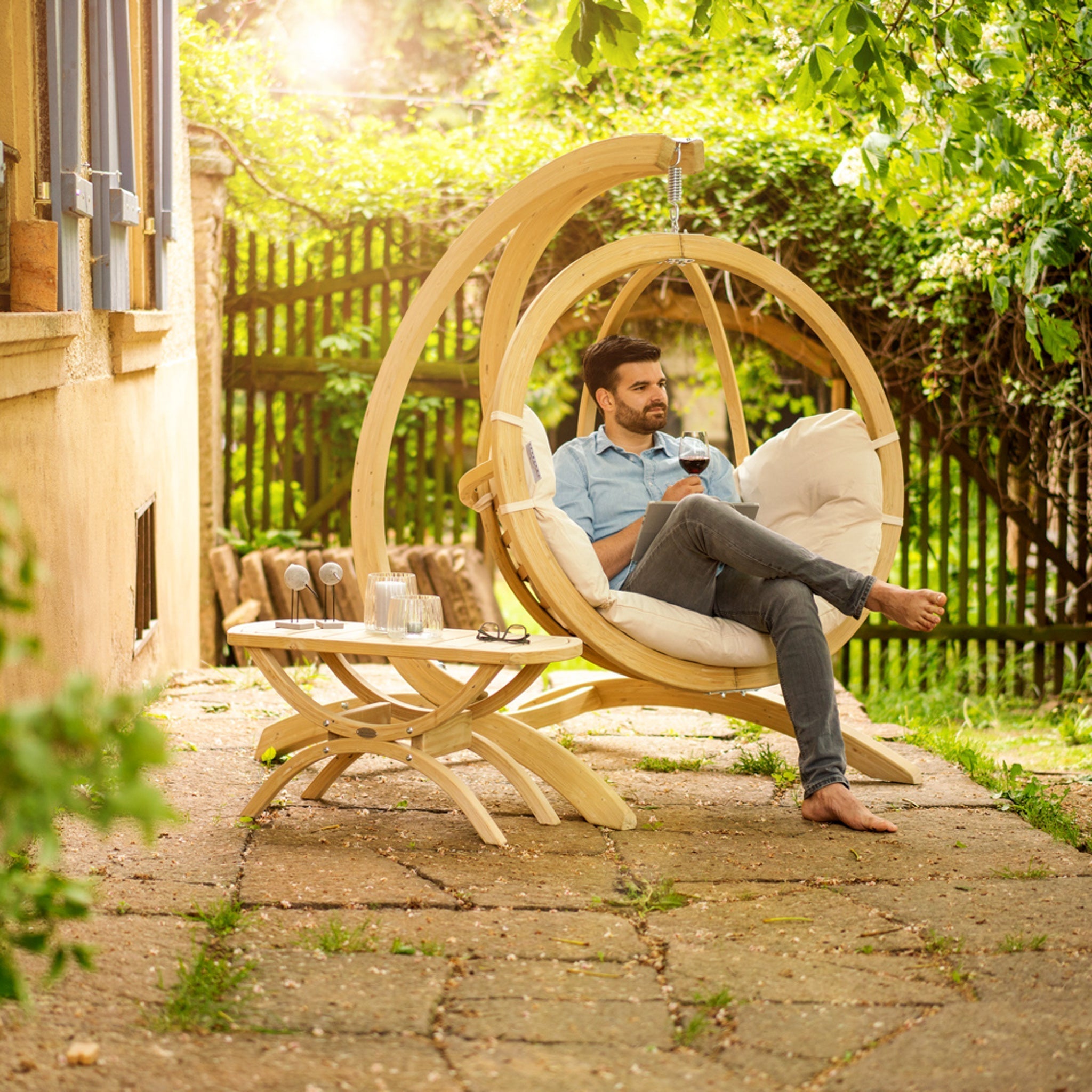 Man relaxing with a glass of wine in the cream beige Globo Hammock Single Seater Chair Set, which features washable cushions, on a sunny patio next to a small table.
