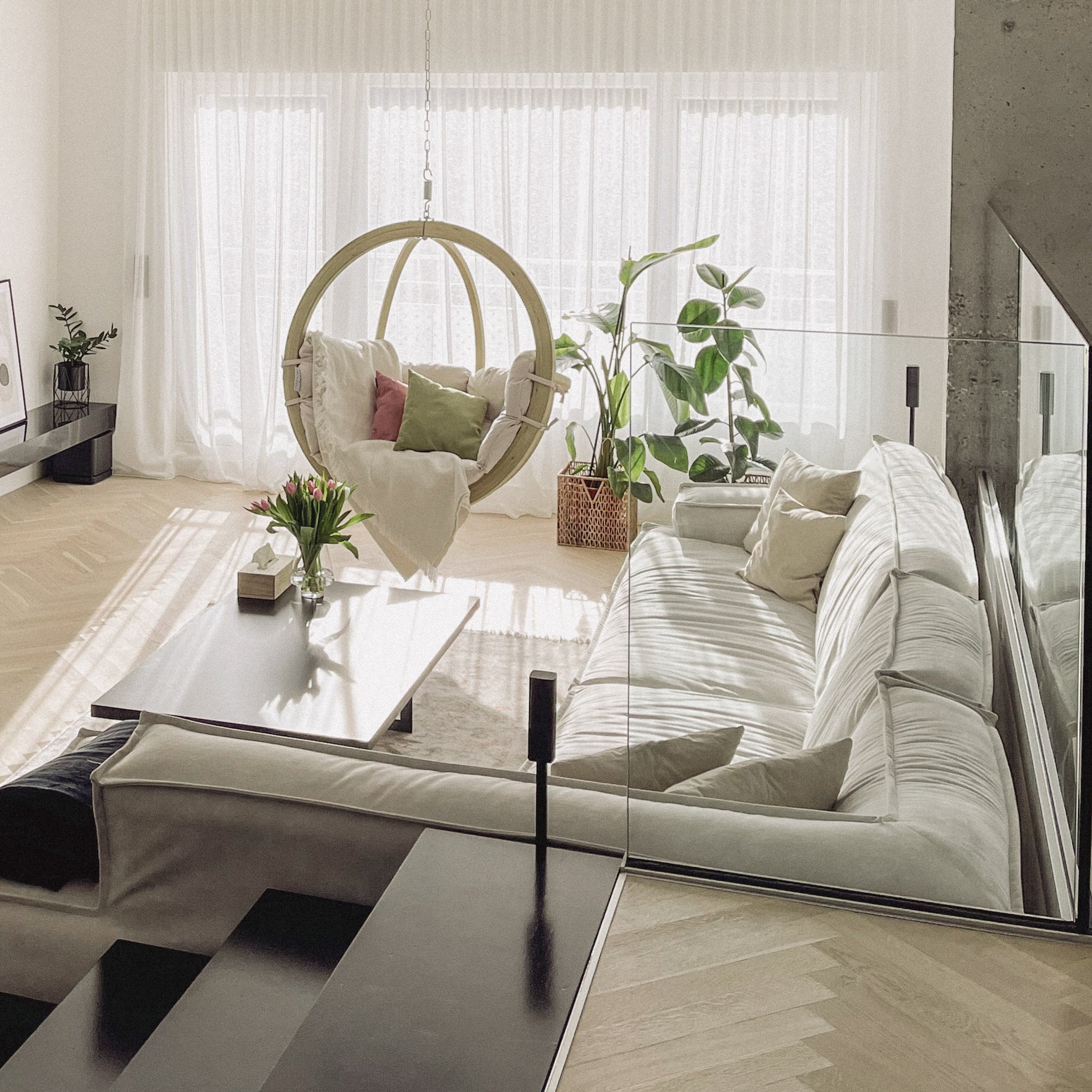 A bright, modern living room with stylish furniture, including a white sofa and comfortable chair. It boasts large windows, plants, the elegant Natura Globo Single Hanging Chair, and a glass railing that seamlessly blends indoor and outdoor living spaces.