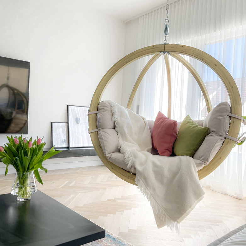 Modern living room with stylish furniture, featuring the Natura Globo Single Hanging Chair, comfortable throw pillows, a vase of tulips on a coffee table, and large windows that blend indoor and outdoor living spaces seamlessly.