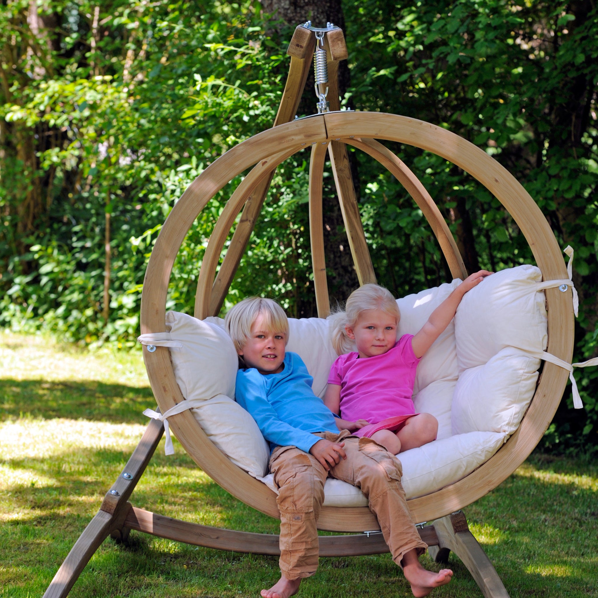 Two children sit together in a stylish Natura Globo Single Hanging Chair outdoors, surrounded by greenery.
