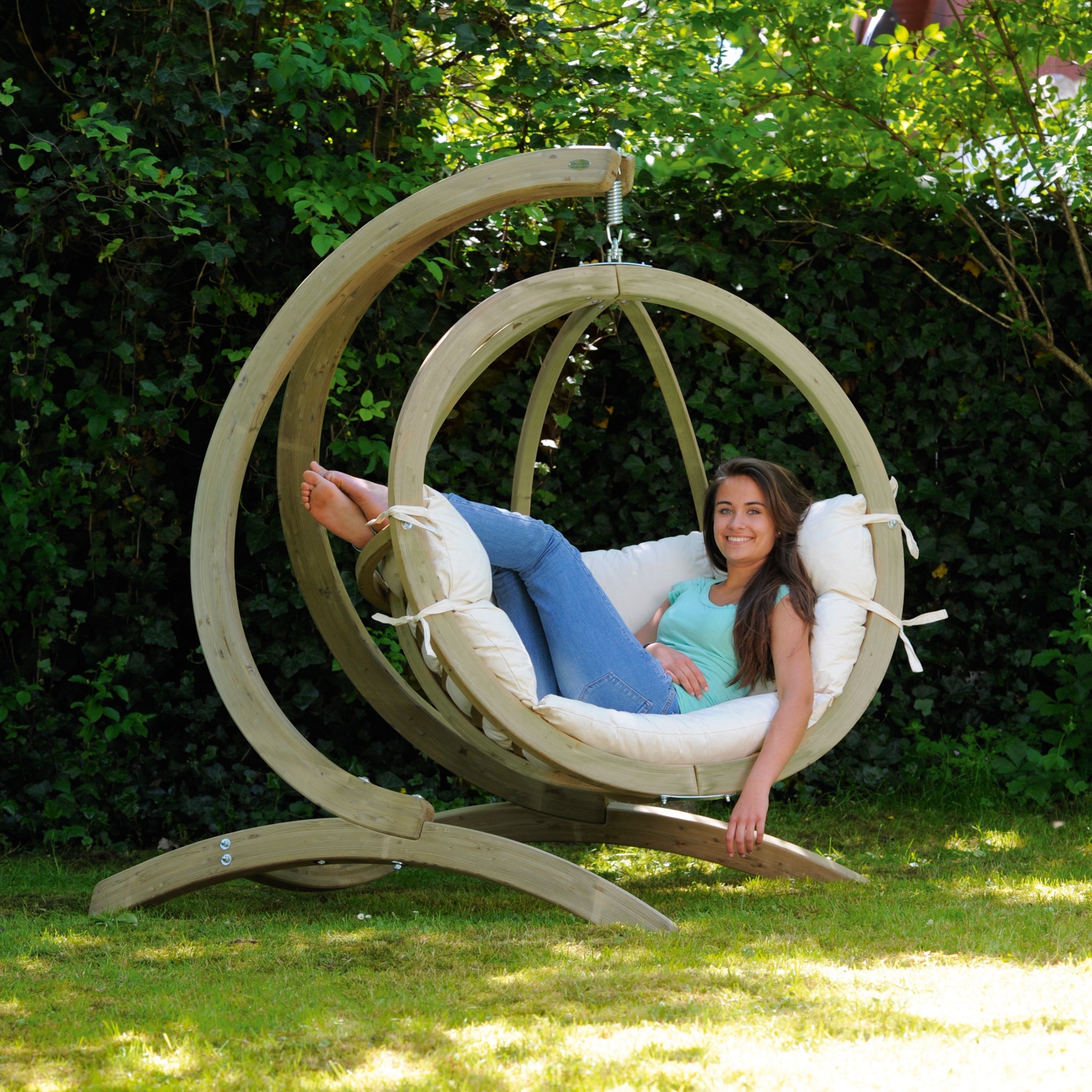 A woman relaxes on the Globo Hammock Single Seater Chair Set in Cream Beige, smiling at the camera while surrounded by weatherproofed spruce in a lush garden and enjoying the comfort of washable cushions.