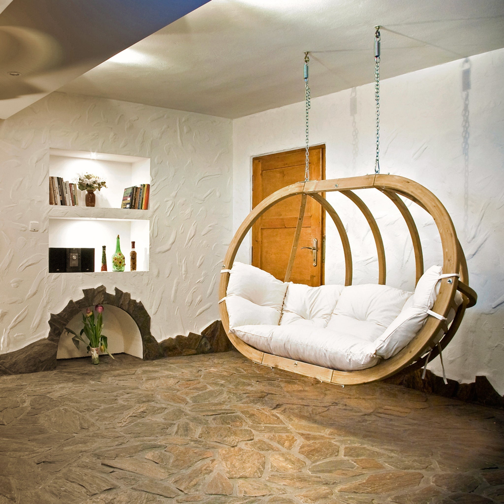 A cozy room with a Globo Royal Double Seater Hanging Chair in Natura, stone floor, wooden door, and recessed shelves holding books and decor. The hanging chair is crafted from weatherproof spruce wood, making it ideal for outdoor use as well.