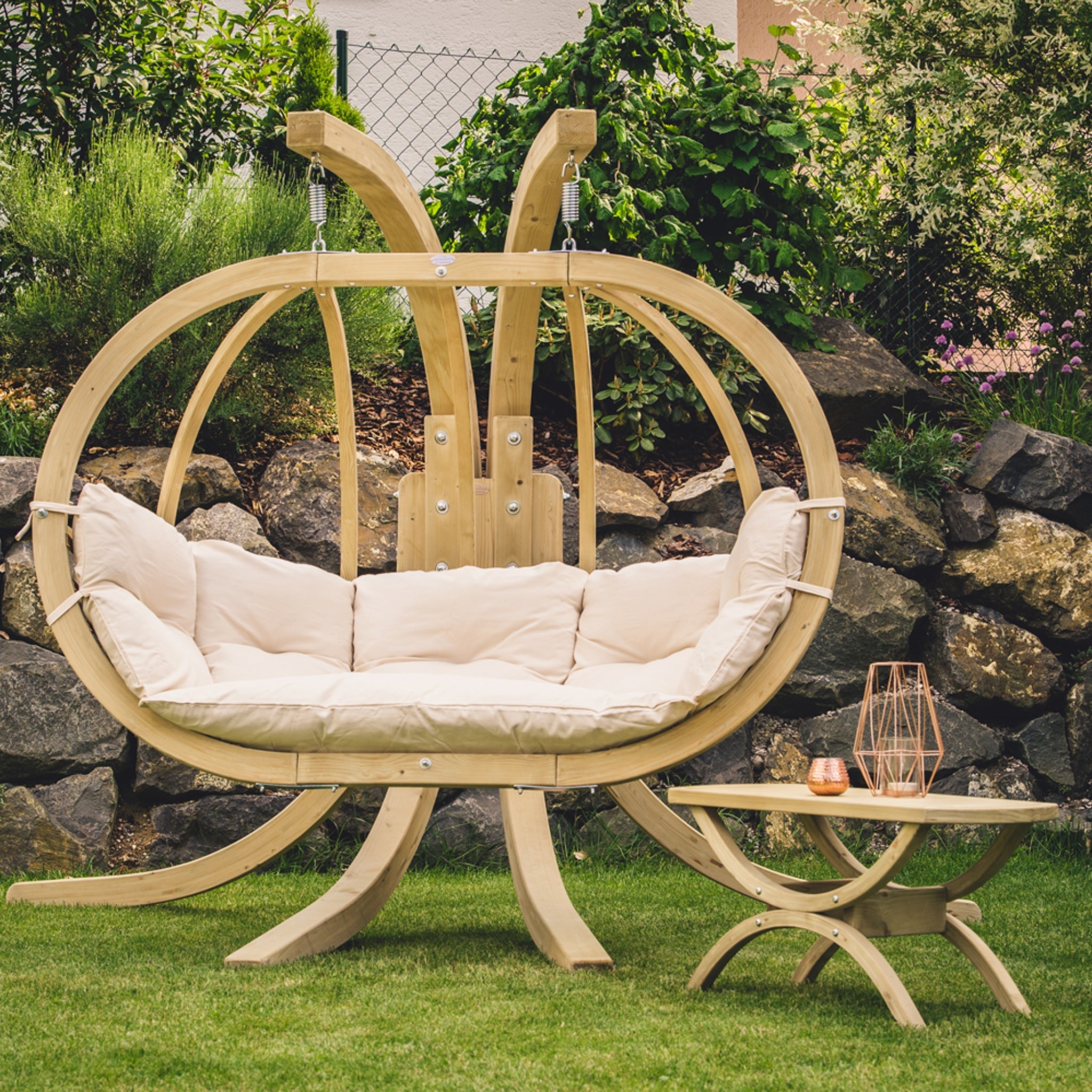 The Globo Royal Double Seater Hanging Chair in Natura, featuring beige cushions and crafted from weatherproof spruce wood for outdoor use, is paired with a small matching wooden table. Set on a grassy lawn with a stone backdrop, it offers an unparalleled relaxation experience just like the renowned comfort of the Globo Royal Double Hammock.