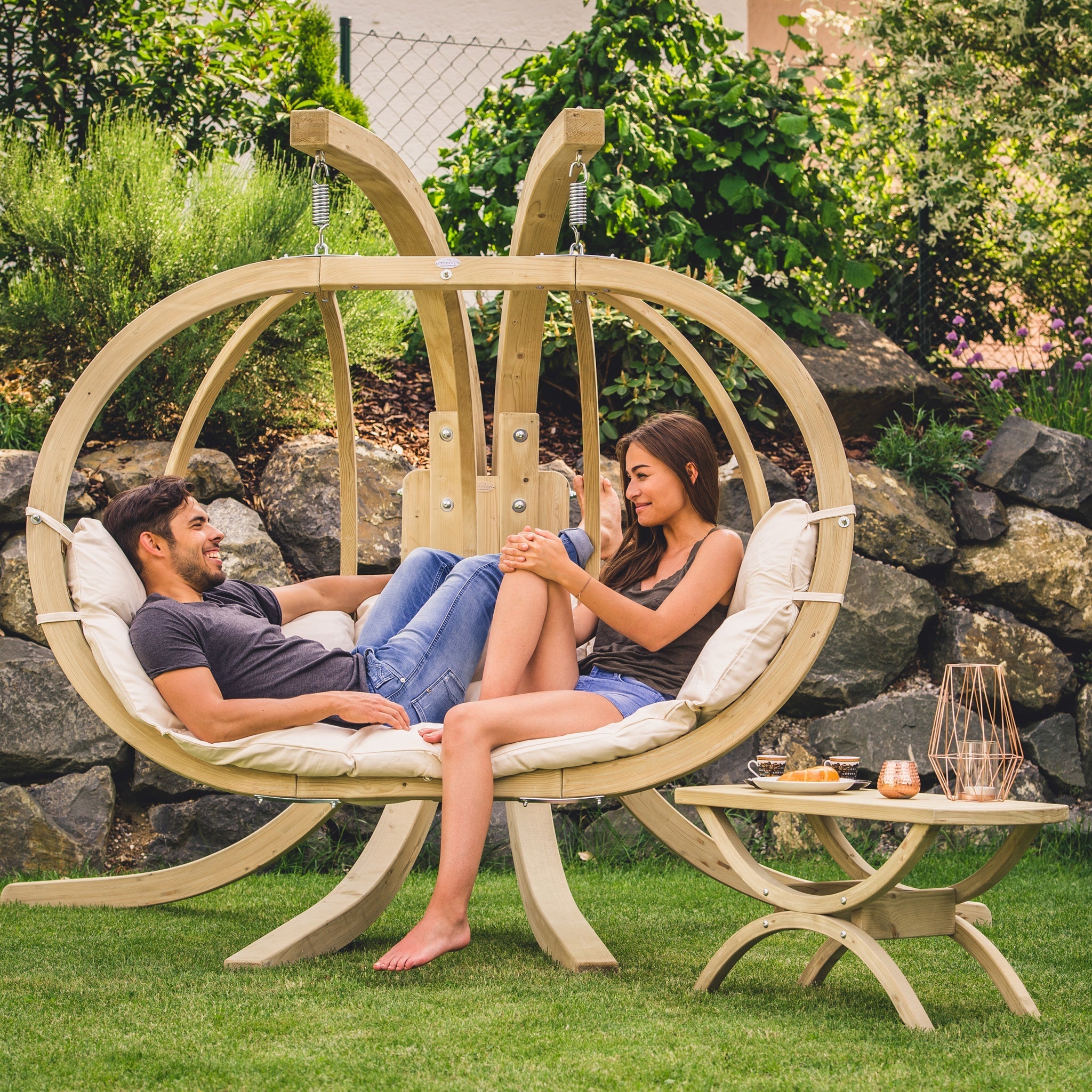 A couple relaxes in a Globo Royal Double Seater Hanging Chair in Natura, swinging gently in a garden. The weatherproof spruce wood ensures it's perfect for outdoor use, with a table holding drinks nearby.