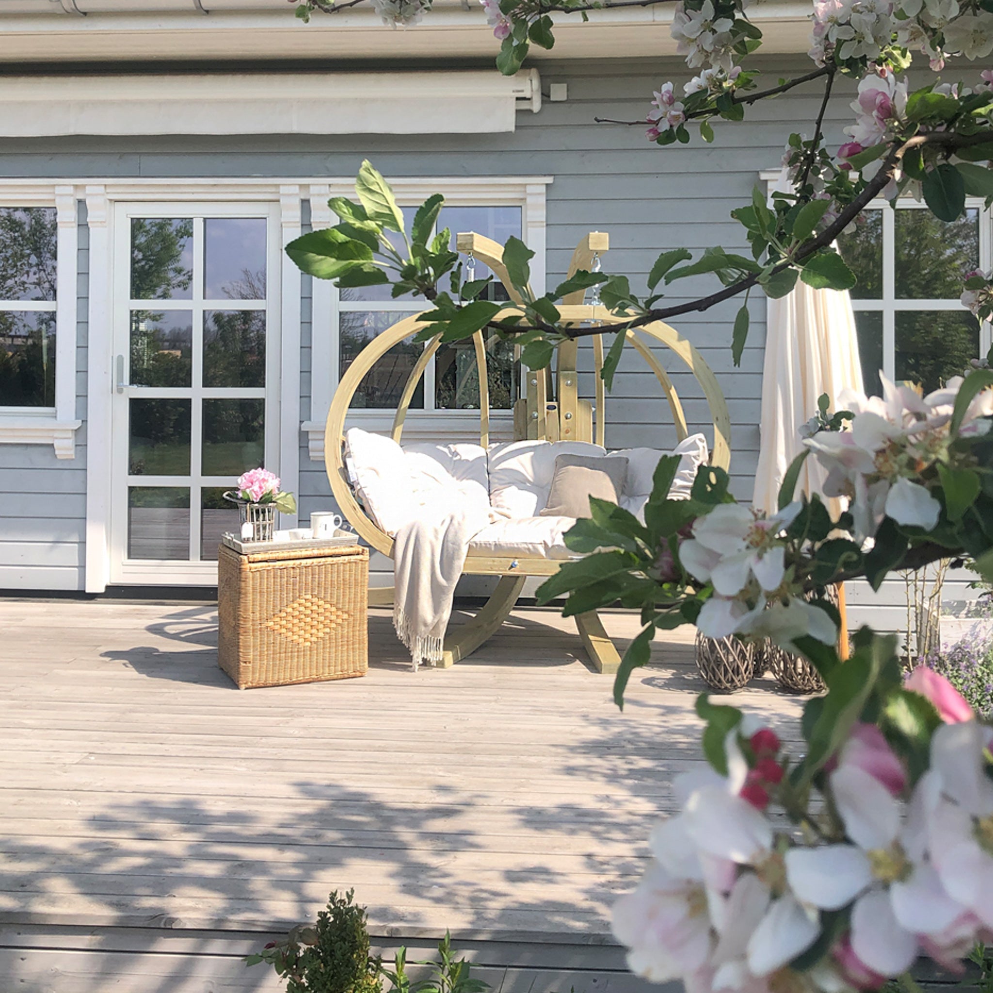 Outdoor deck with a cozy Globo Royal Double Seater Hanging Chair in Natura, a wicker side table, and blossoming tree branches in the foreground. Ideal for outdoor use, the hanging chair is crafted from weatherproof spruce wood for lasting durability.
