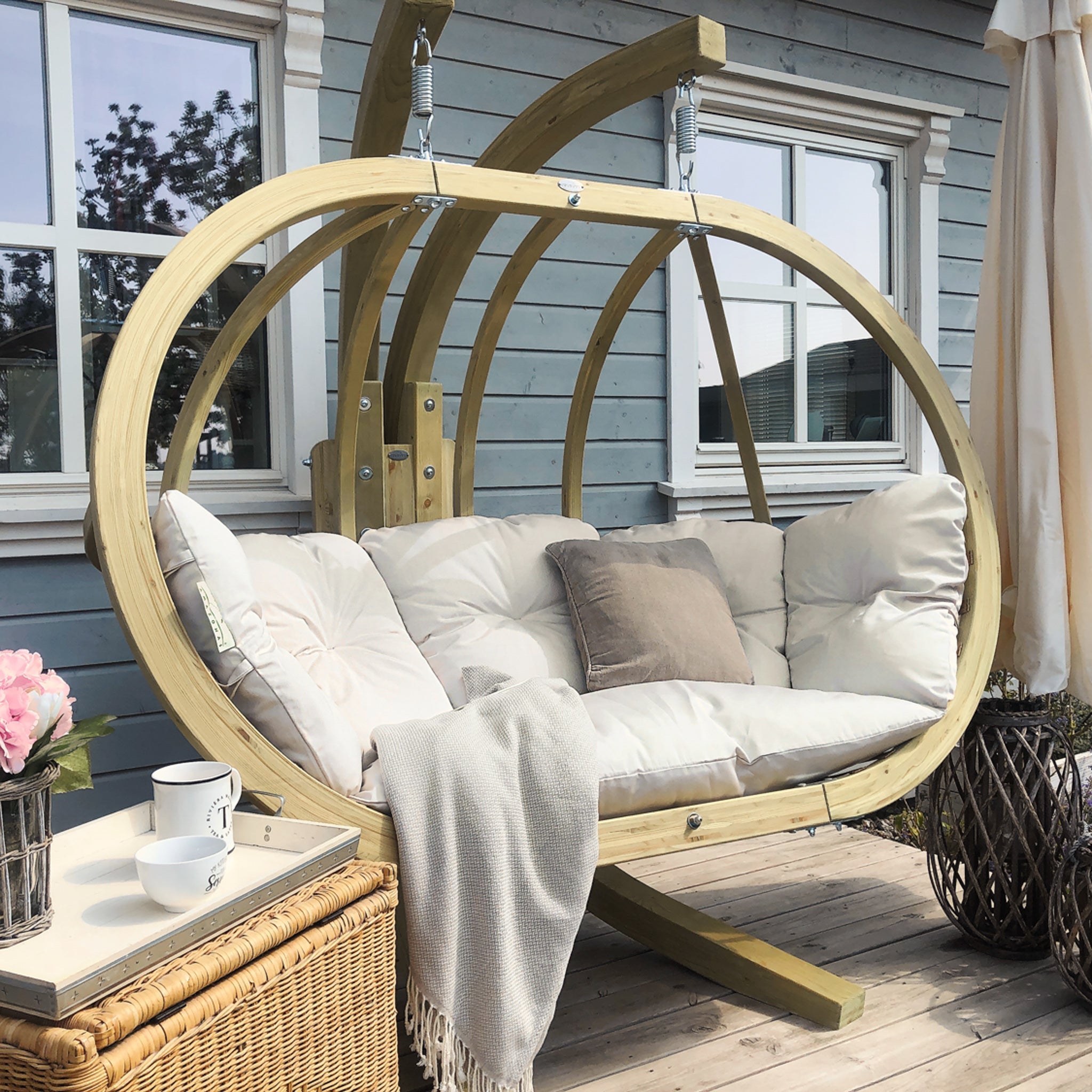 A weatherproof Globo Royal Double Seater Hanging Chair in Natura with cushions on a patio, accompanied by a table, a cup, a vase, and a blanket nearby. Perfect for outdoor use, this hanging chair offers both comfort and durability.