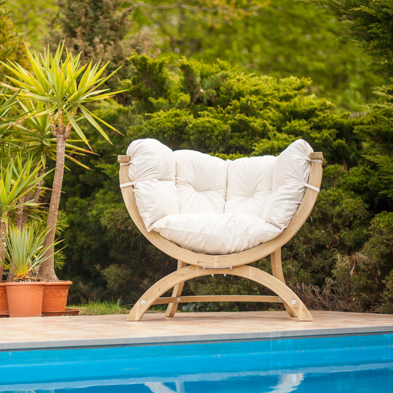 A round Globo Siena Uno Seat in Cream Beige with an upholstered washable cushion sits by a pool, enveloped by potted plants and lush greenery. Crafted from sustainably sourced wood, this chair effortlessly merges comfort with eco-conscious design.