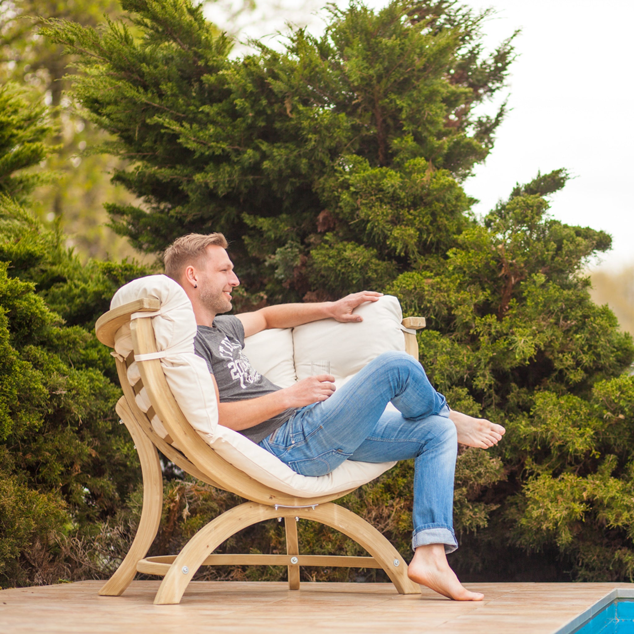 A man sits comfortably in a Globo Siena Uno Seat in Cream Beige by a pool, smiling and wearing jeans and a t-shirt, the sustainably sourced wood frame complemented by an upholstered washable cushion.