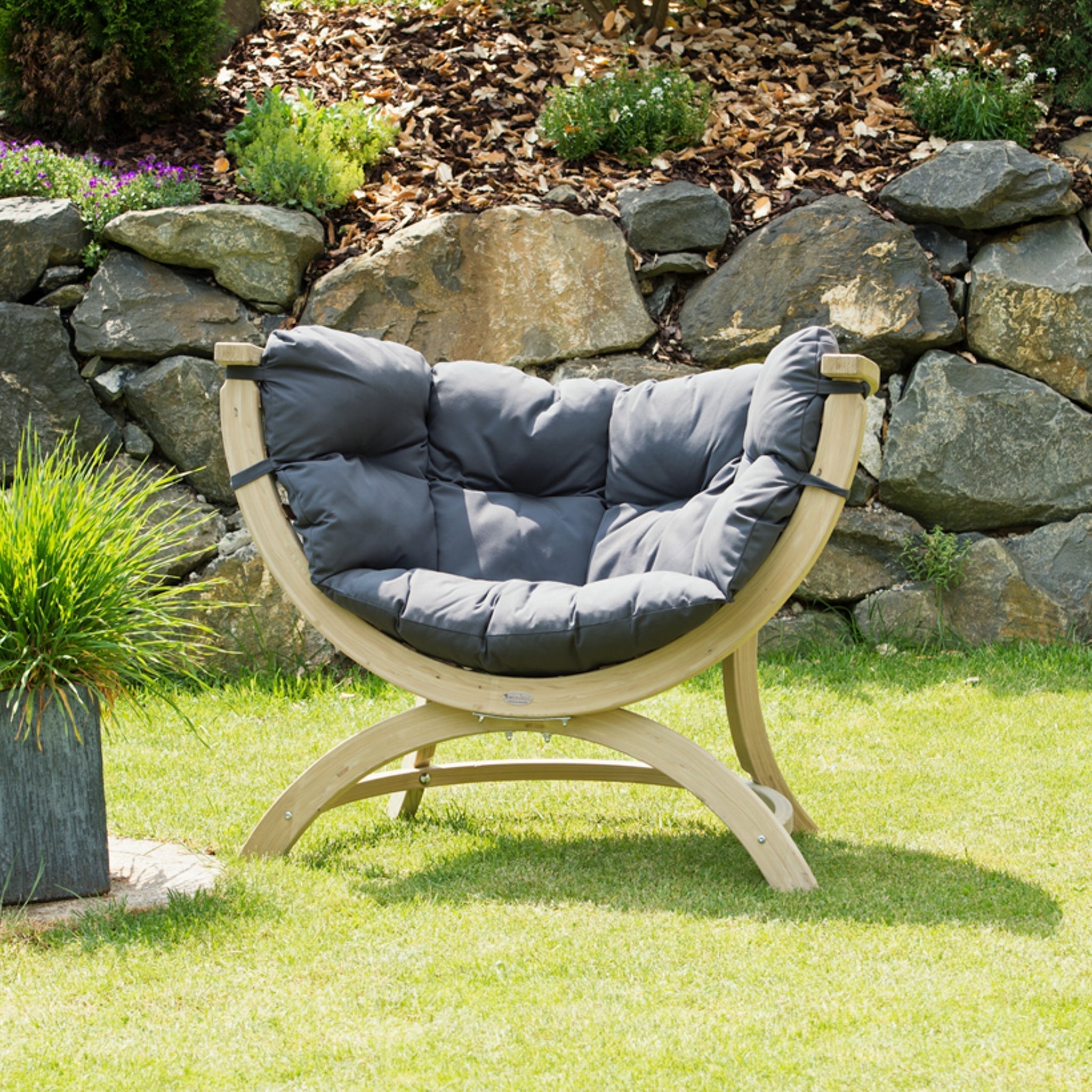 A Globo Siena Uno Seat Weatherproof in Anthracite Grey rests on a green lawn, with a stone wall and plants in the background.