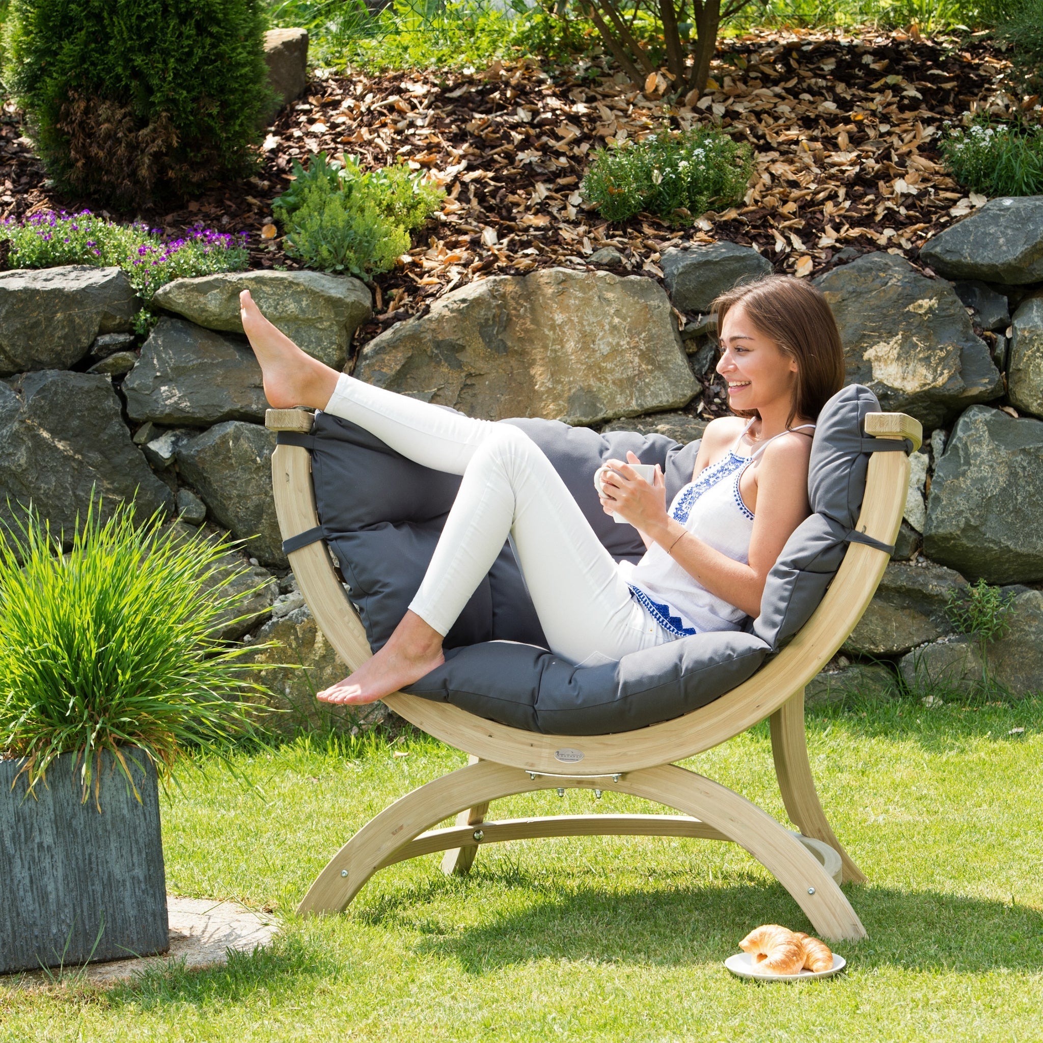 A woman relaxes on the Globo Siena Uno Seat Weatherproof in Anthracite Grey, a wooden chair crafted from sustainably sourced wood, holding a cup with a pastry nearby on the ground.