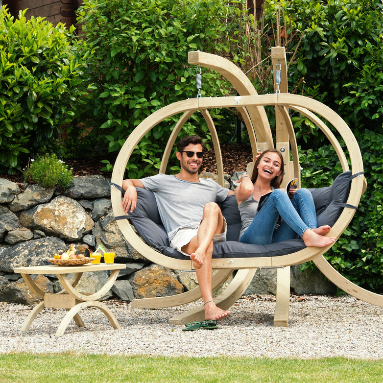 Two people relax outdoors on the Globo Royal Double Seater Hanging Chair Set, made from weatherproof spruce wood with anthracite grey galvanised metal fixings. They smile with drinks on a small table nearby, enjoying the stylish and durable setup.