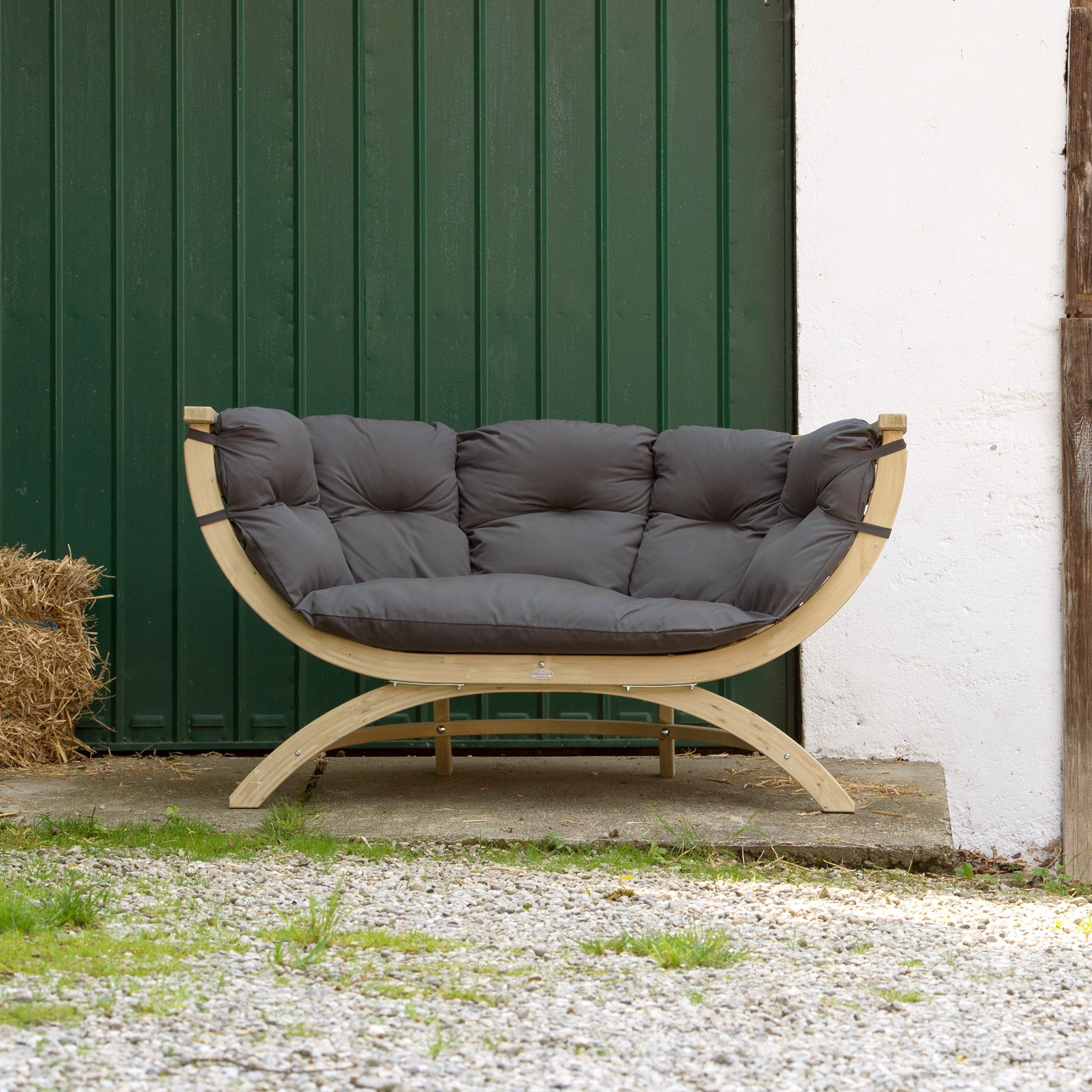 A cushioned wooden bench, the Globo Siena Due Seat Weatherproof in Anthracite Grey, crafted from sustainably sourced wood, is placed outdoors near a green door and some hay bales.