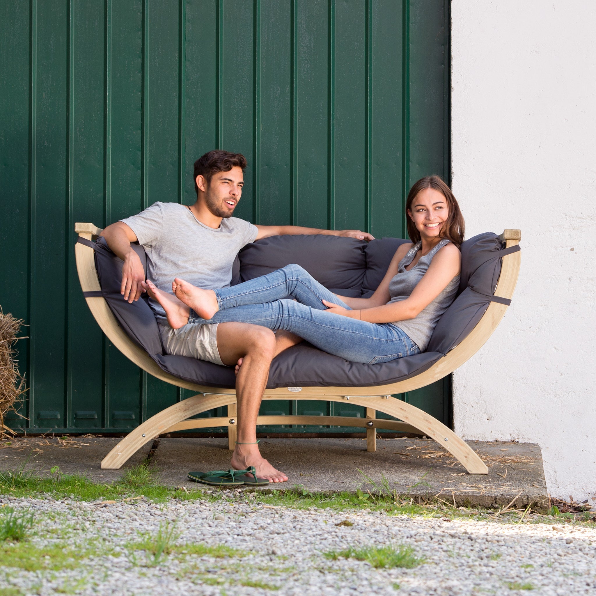 Two people sit and smile on the Globo Siena Due Seat Weatherproof in Anthracite Grey, a cushioned wooden outdoor seat crafted from sustainably sourced wood, in front of a large green door.