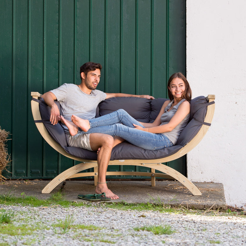 A man and woman relax, smiling and talking, on the Globo Siena Due Seat Weatherproof in Anthracite Grey made from sustainable wood.