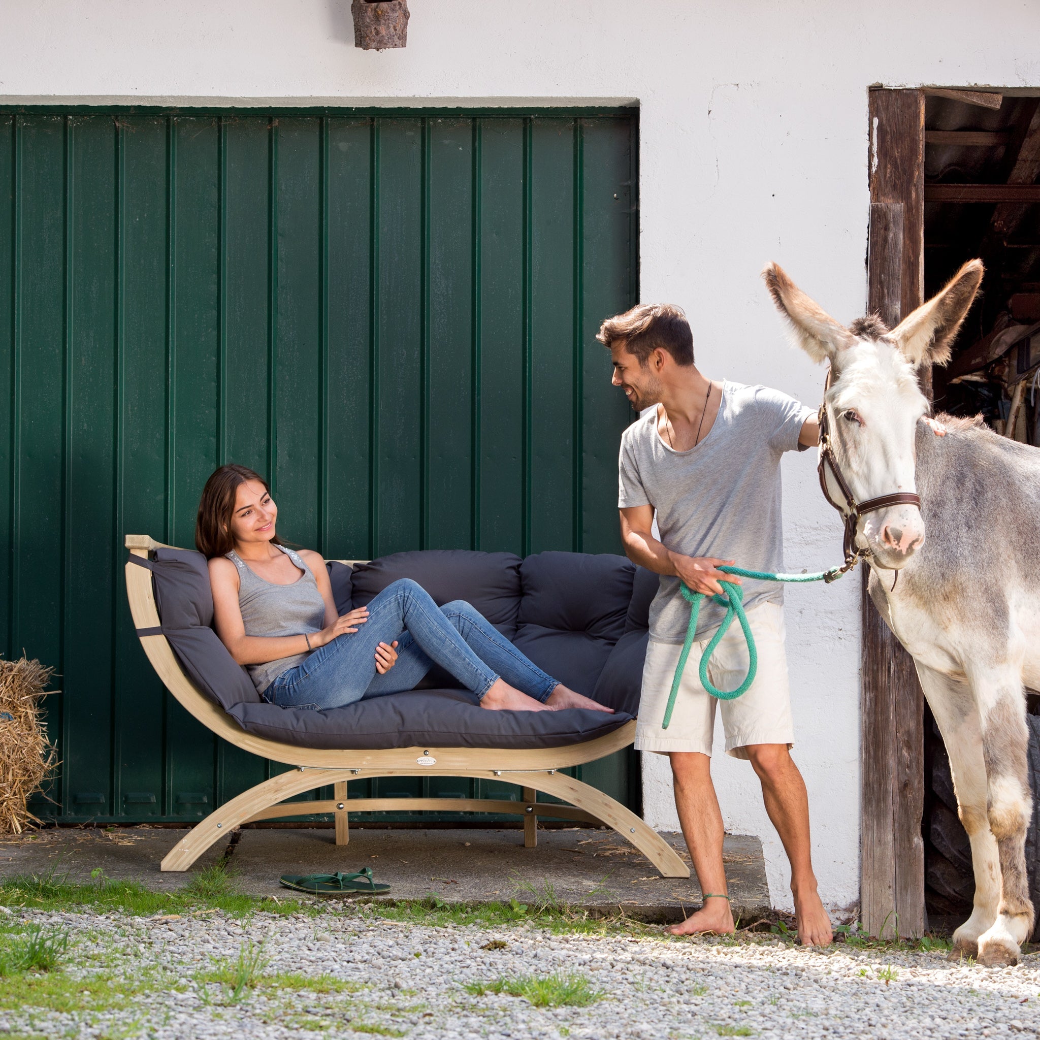 Woman relaxing on a Globo Siena Due Seat Weatherproof in Anthracite Grey while a man stands next to her, holding a donkey by a green leash outside a building.