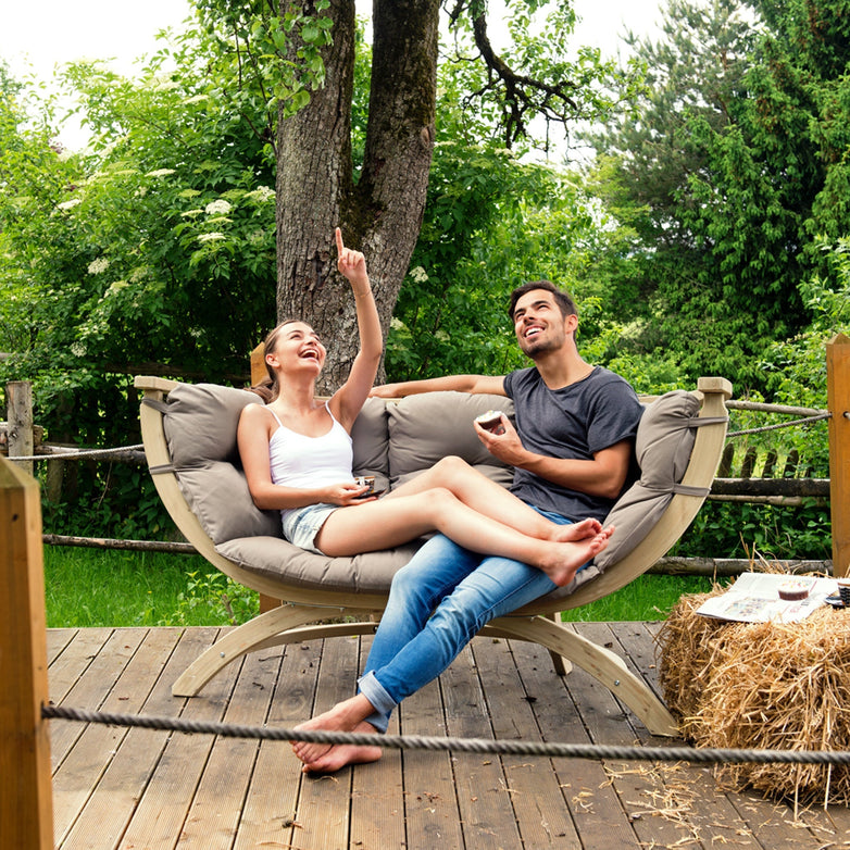 A smiling couple relaxes on the Globo Siena Due Seat Weatherproof in Taupe Silver, laughing and looking up while surrounded by lush greenery.