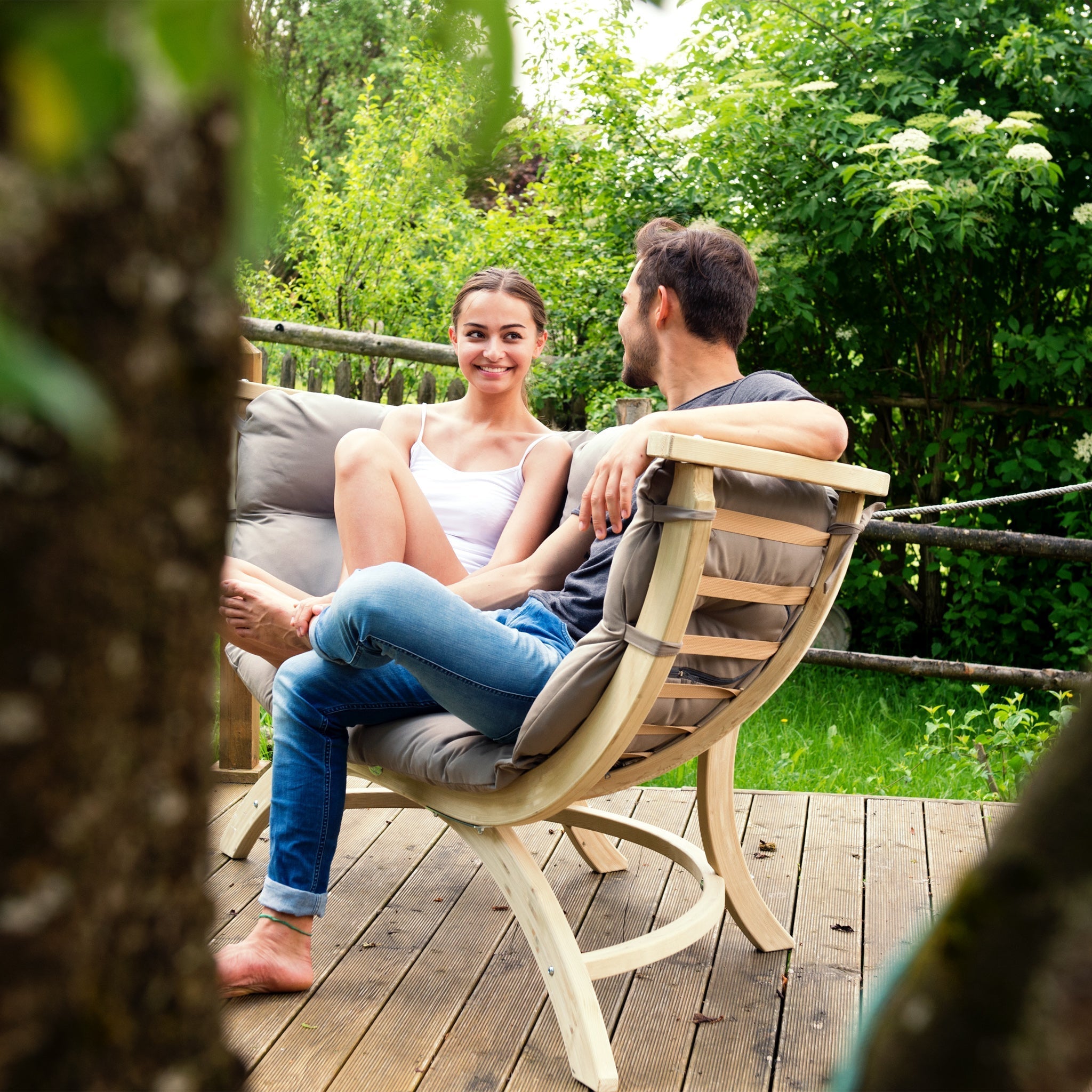 A woman and a man sit on the elegant Globo Siena Due Seat Weatherproof in Taupe Silver on a wooden deck, surrounded by lush greenery, smiling and talking. The sustainable wood of the deck enhances the serene atmosphere, making it perfect for both outdoor and indoor use.