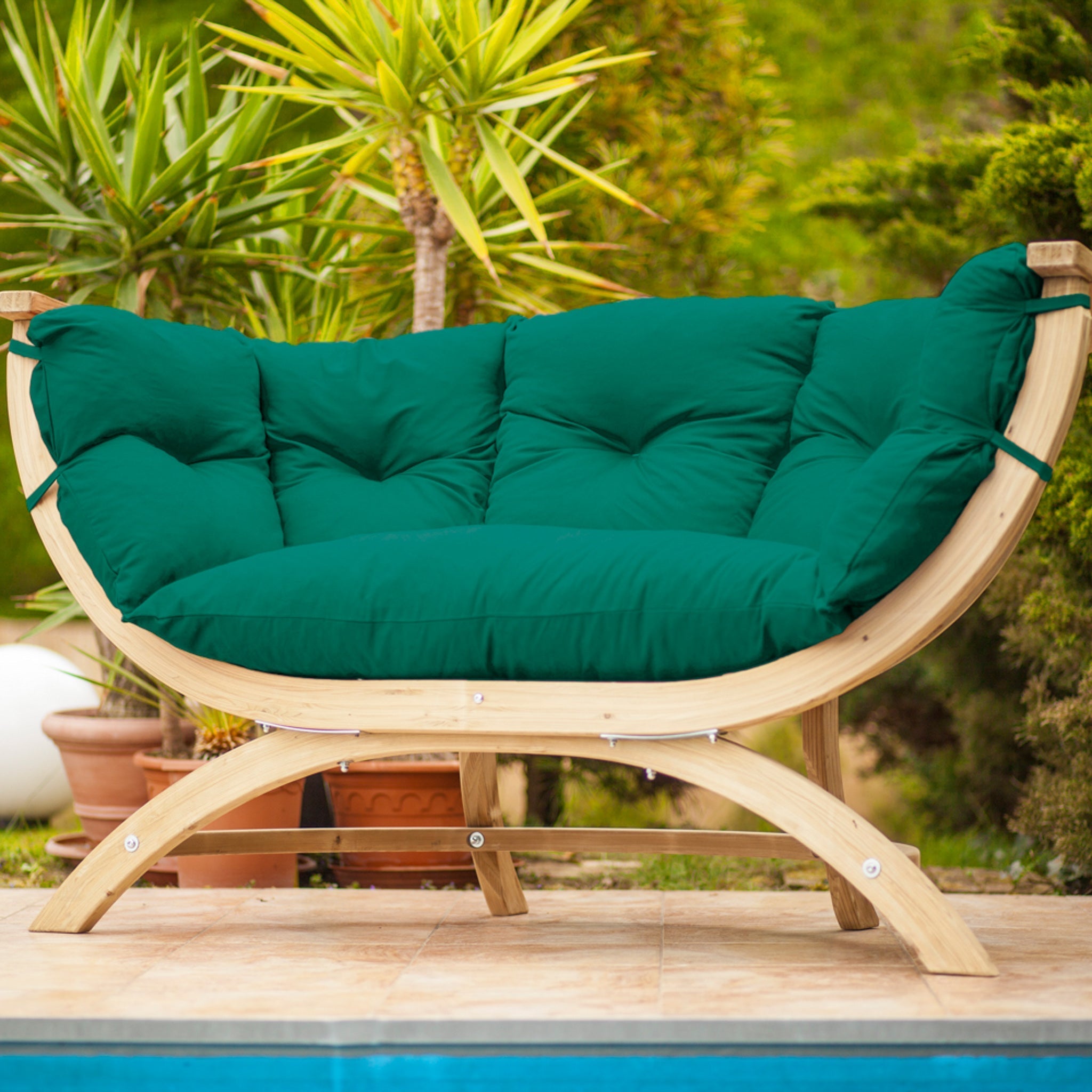 The Globo Siena Due Seat Weatherproof in Verde Green is an elegant seating option, set beside a pool and surrounded by lush green tropical plants. Crafted from sustainable wood, this outdoor chair with green, washable cushions seamlessly combines style and eco-friendliness.