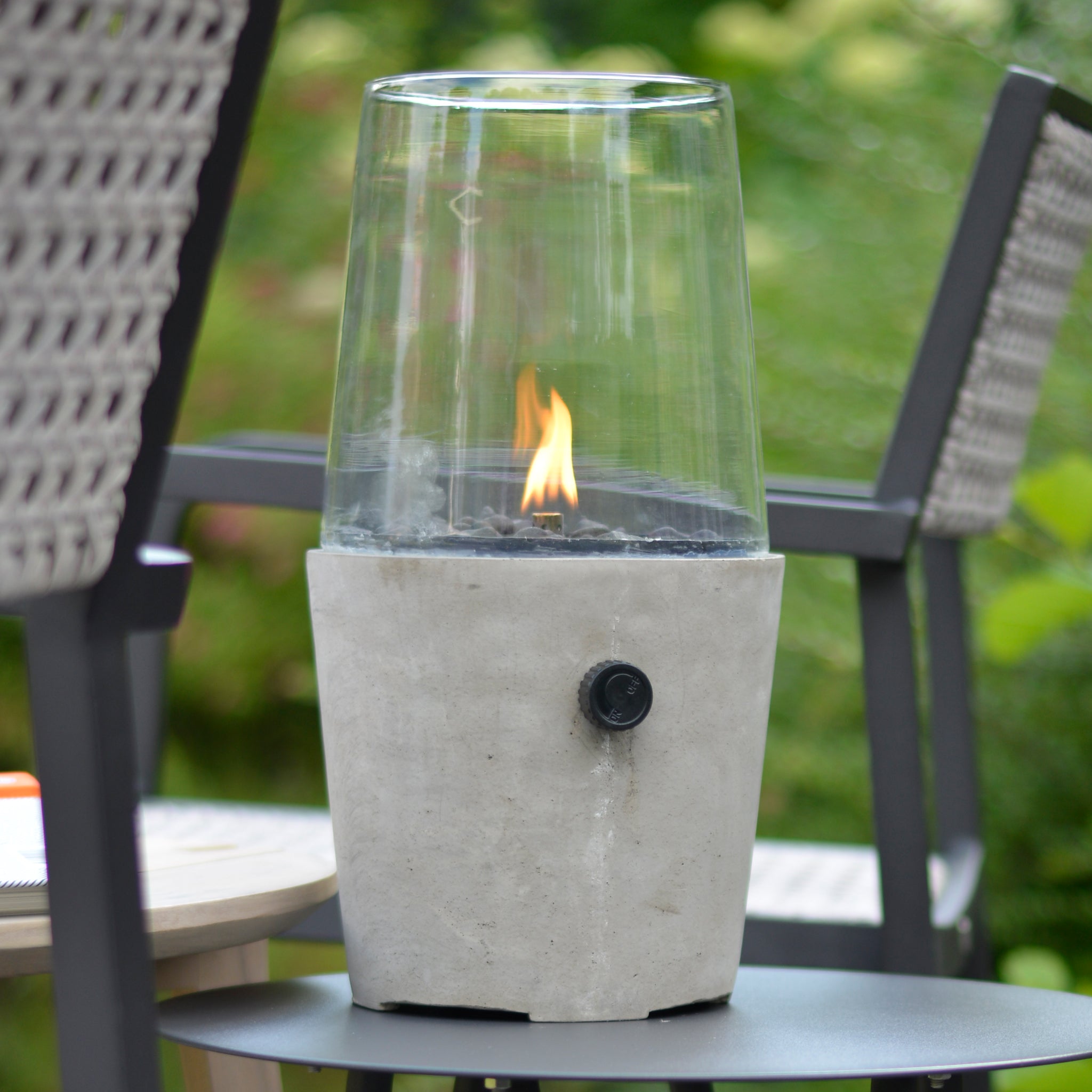 The Cosi - Cosicement Round Fire Lantern, featuring a glass-enclosed tabletop design with a stylish concrete finish, is perfectly suited to be surrounded by outdoor chairs.