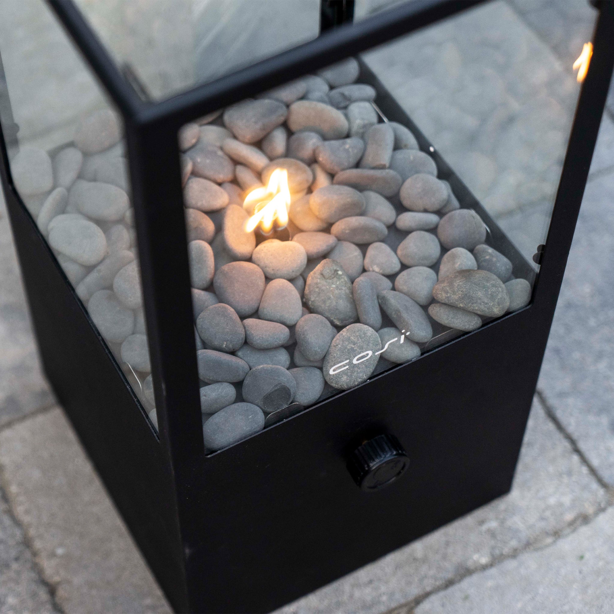 The Cosi Cosidome High Black Fire Lantern features a minimalistic design with a small flickering flame inside a glass casing, surrounded by gray pebbles on a stone base.