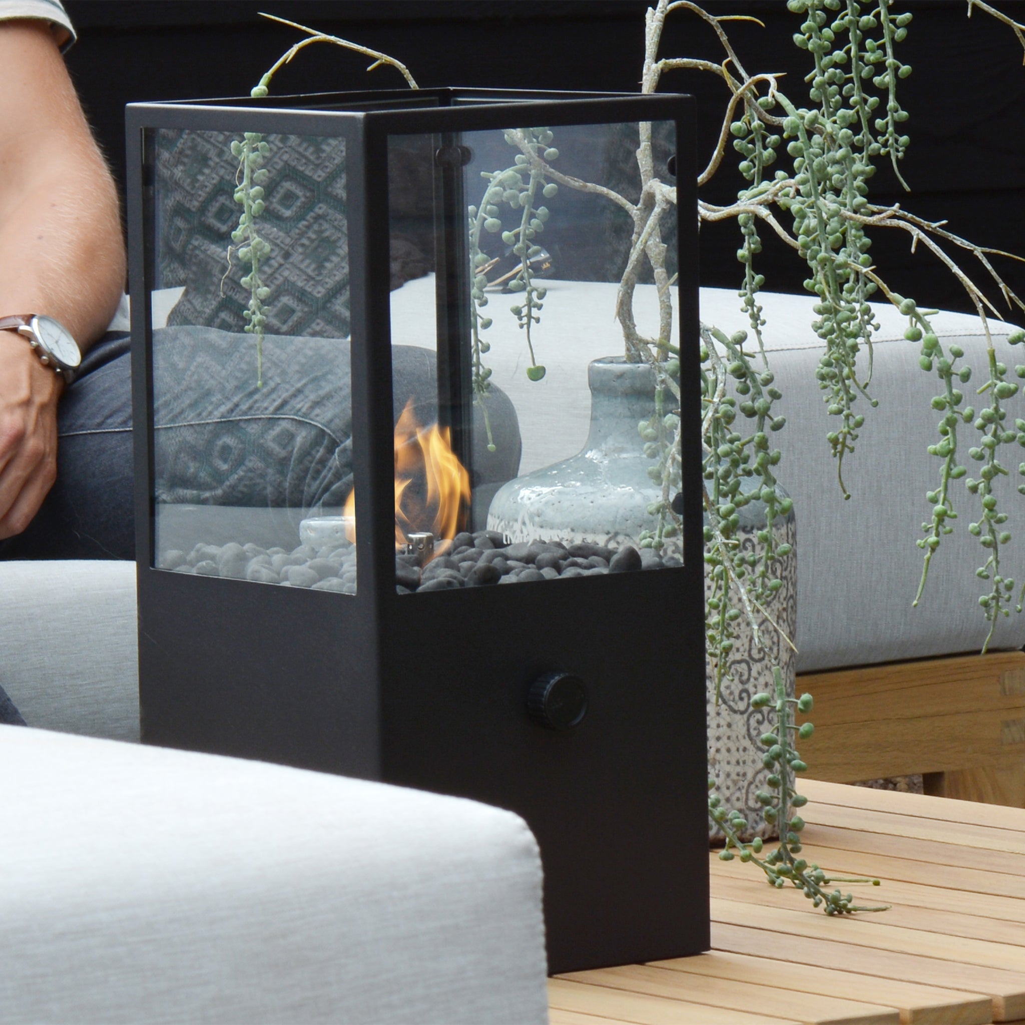 The Cosi - Cosidome High Black Fire Lantern, featuring a sleek glass enclosure, sits on a wooden table surrounded by people and plants. Its minimalistic design enhances elegance and creates a cozy atmosphere for everyone gathered around.