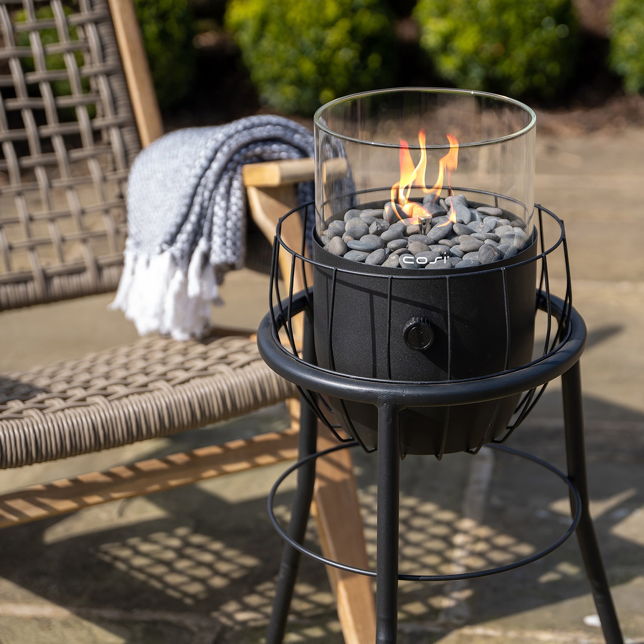 The Cosi - Cosiscoop Basket High Black Lantern, placed beside a wicker chair with a gray blanket, provides a warm glow and modern chic outdoor lighting for cozy evenings.