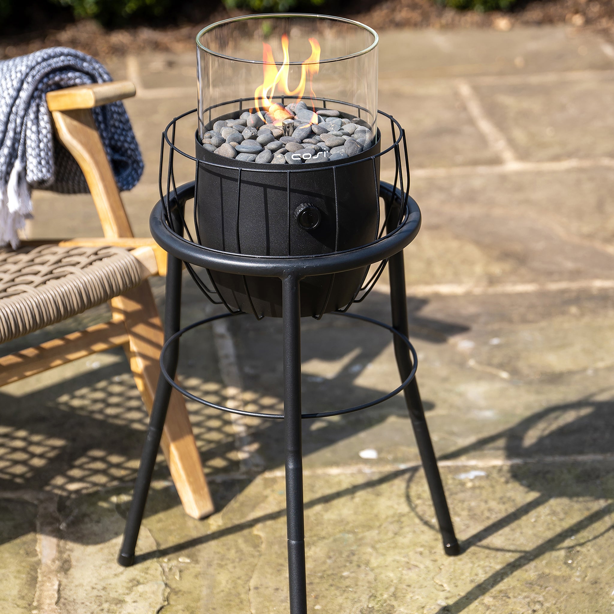 A modern outdoor fire pit with a glass shield and pebble stones is next to a sleek Cosi - Cosiscoop Basket High Black Lantern and a woven chair on a stone patio.