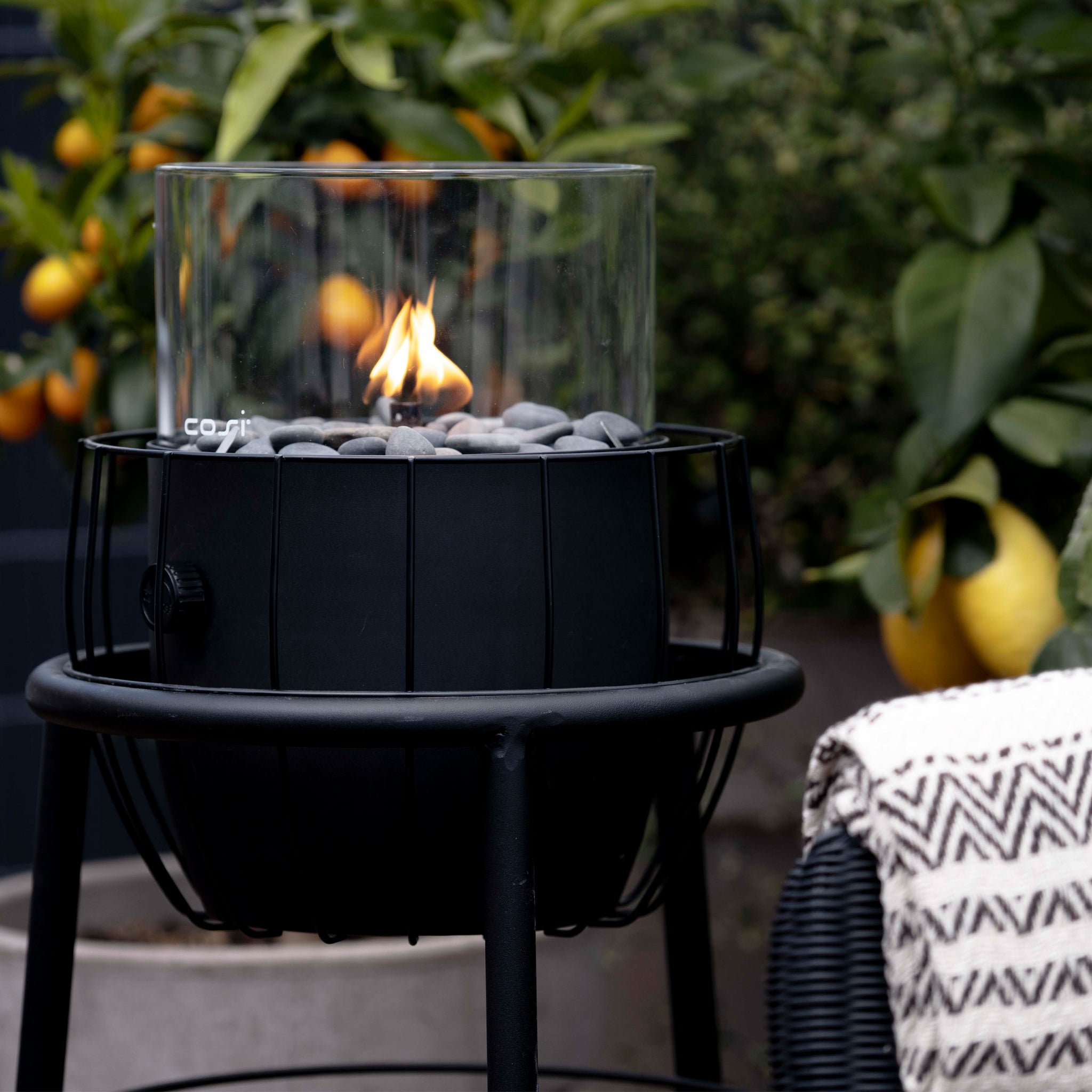 Modern outdoor fire pit with a glass enclosure, surrounded by a lemon tree and enhanced by the chic Cosi - Cosiscoop Basket High Black Lantern. The cozy setting is complete with a blanket draped nearby for added warmth and style.